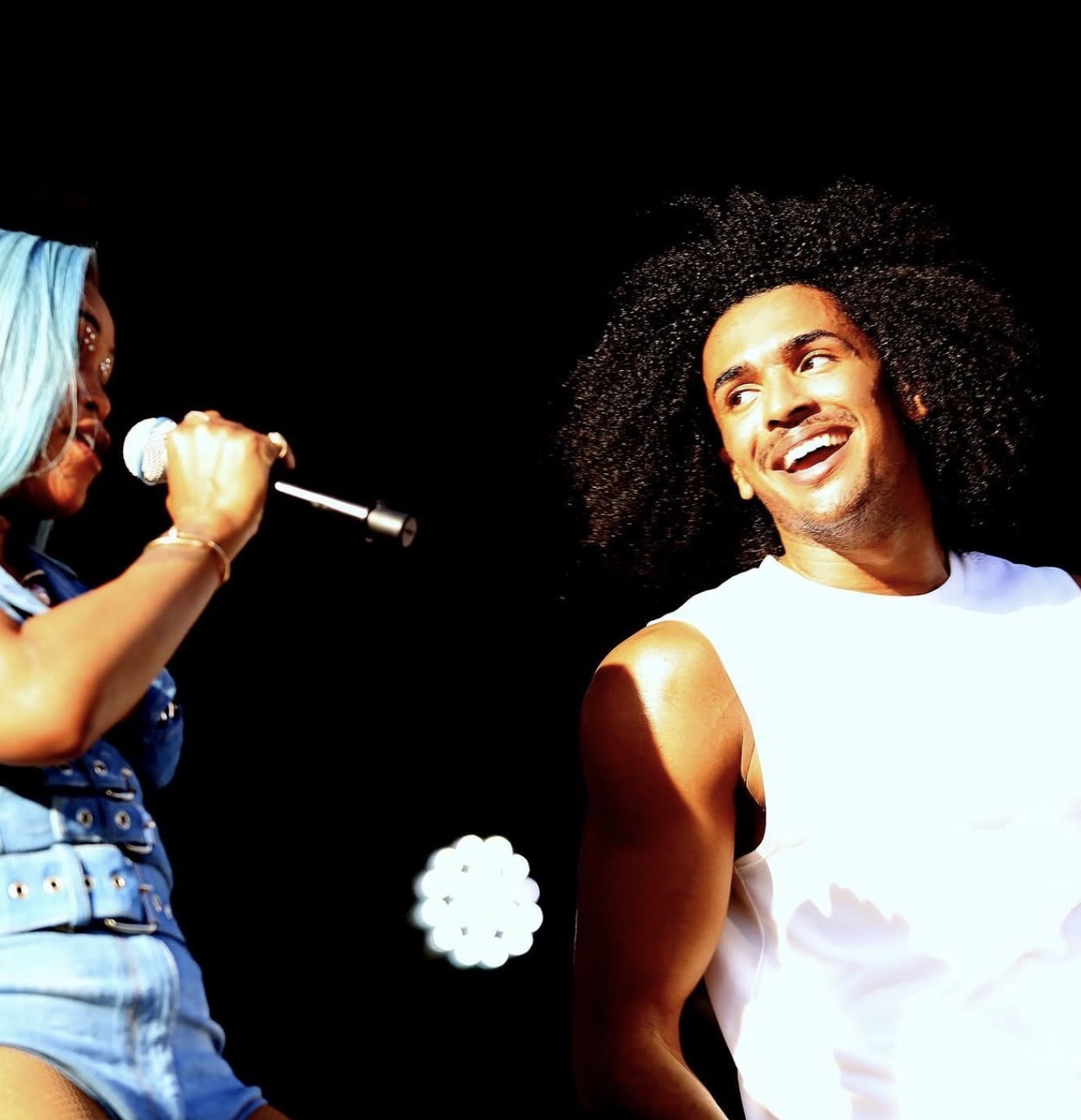 Two people on stage, one holding a microphone, smiling at each other against a black background.