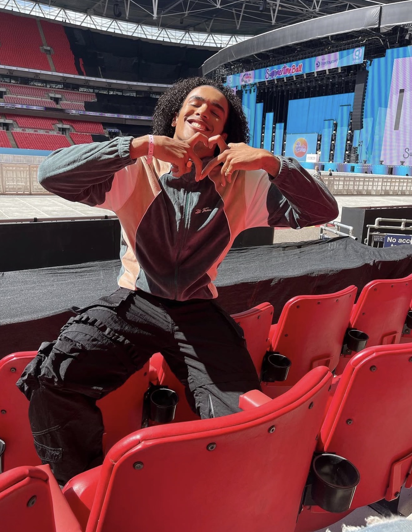Person smiling and making a heart shape with hands in an empty stadium with red seats and a stage in the background.