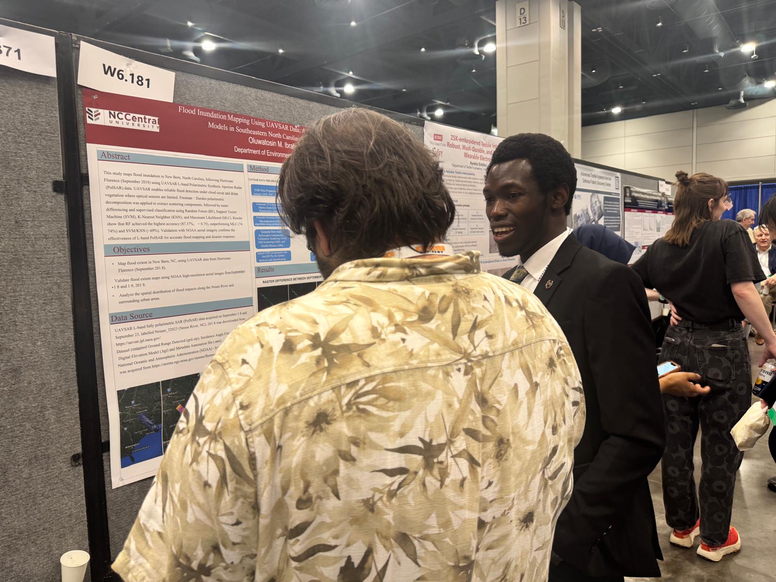Two people in front of a research poster at an indoor conference, one with curly hair and a floral shirt, the other in a suit.