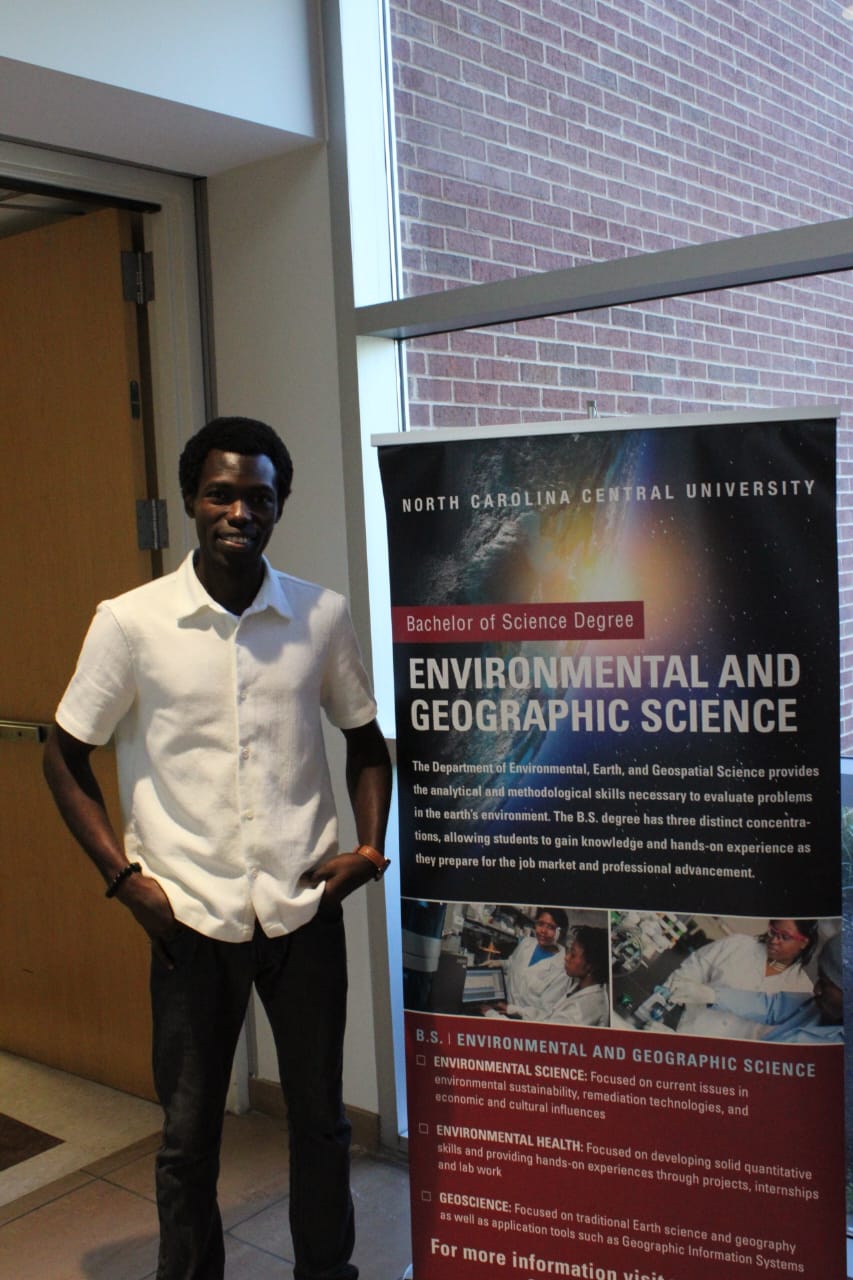 Young man in white shirt standing indoors near a window and a university banner.