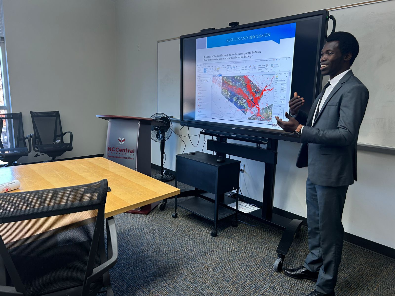 Man in suit giving presentation in conference room with large screen displaying a colorful chart.