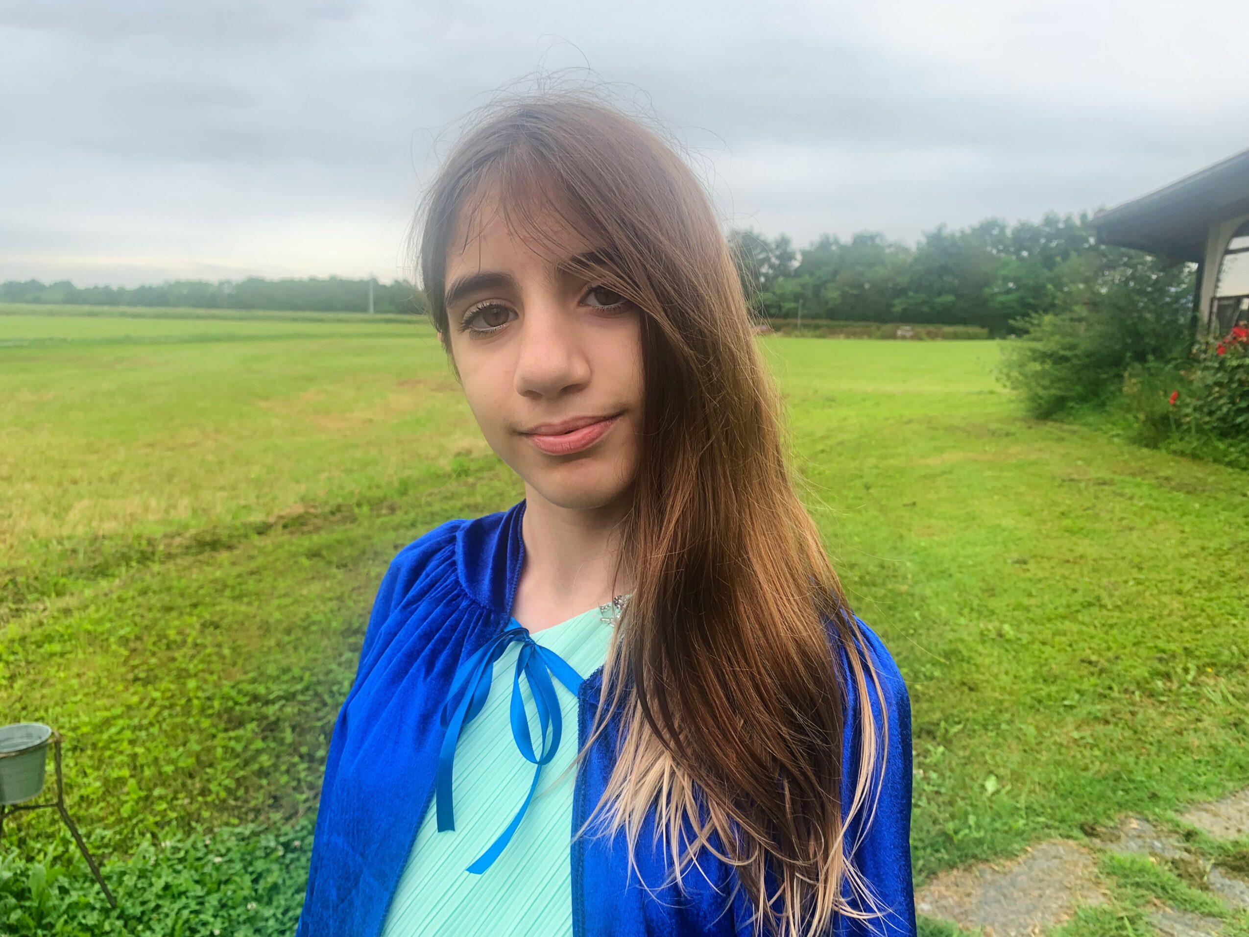 Young girl with long hair standing outdoors on a grassy field, cloudy sky background.