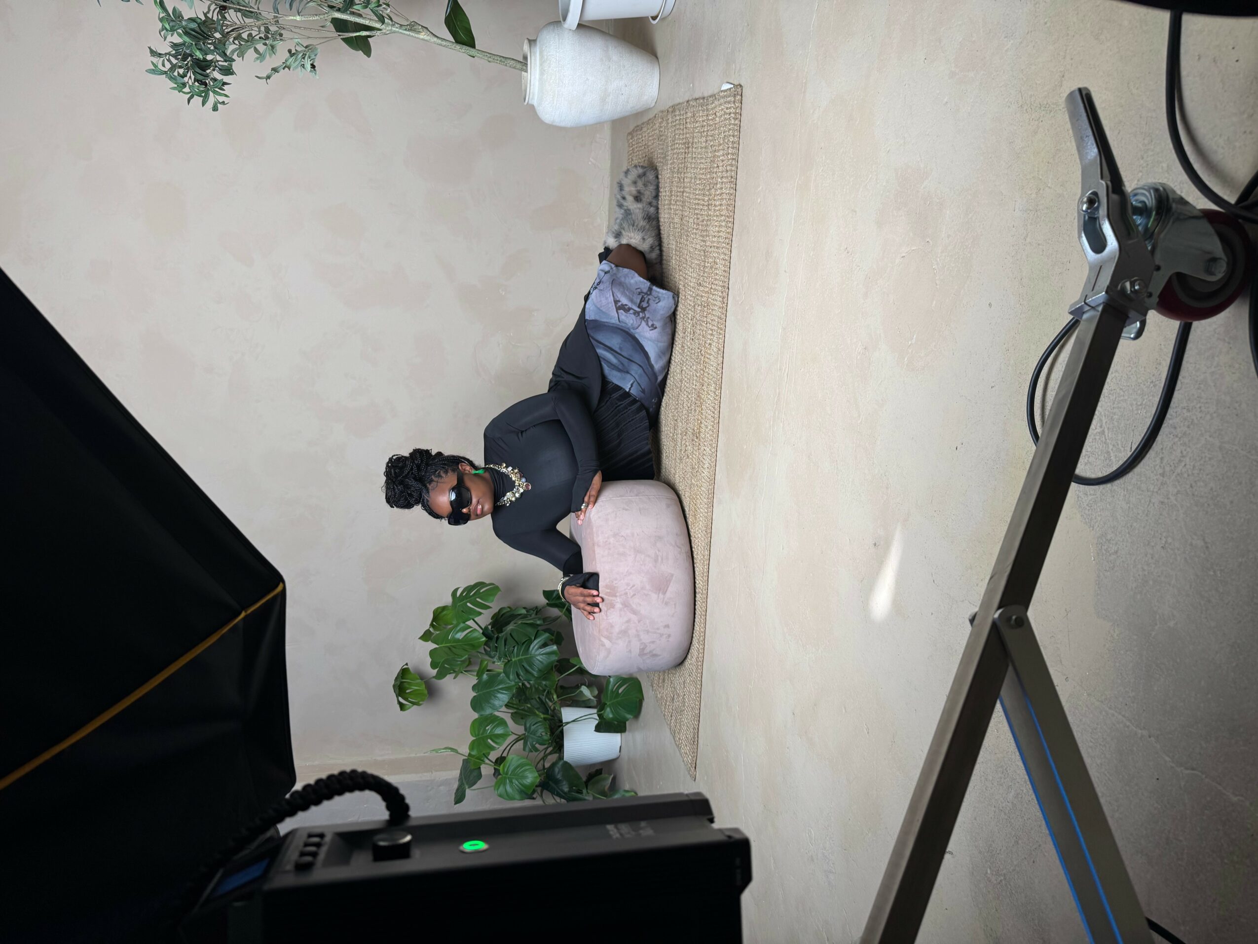 Person lying on floor with a pink cushion, surrounded by plants and a white wall, in a room with a beige floor.