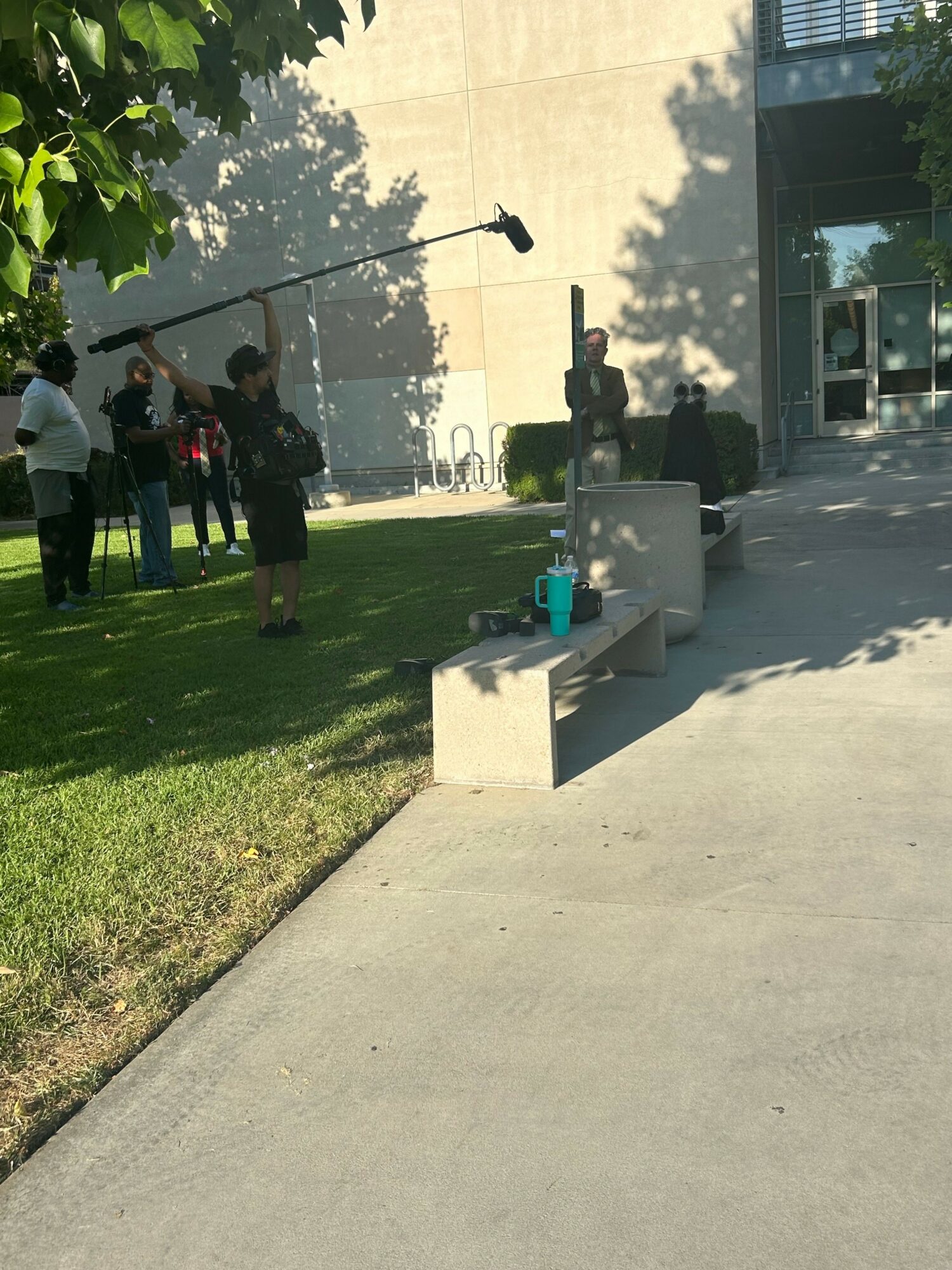 Group of people outdoors near a building, some holding a boom microphone, on a sunny day with shadows.