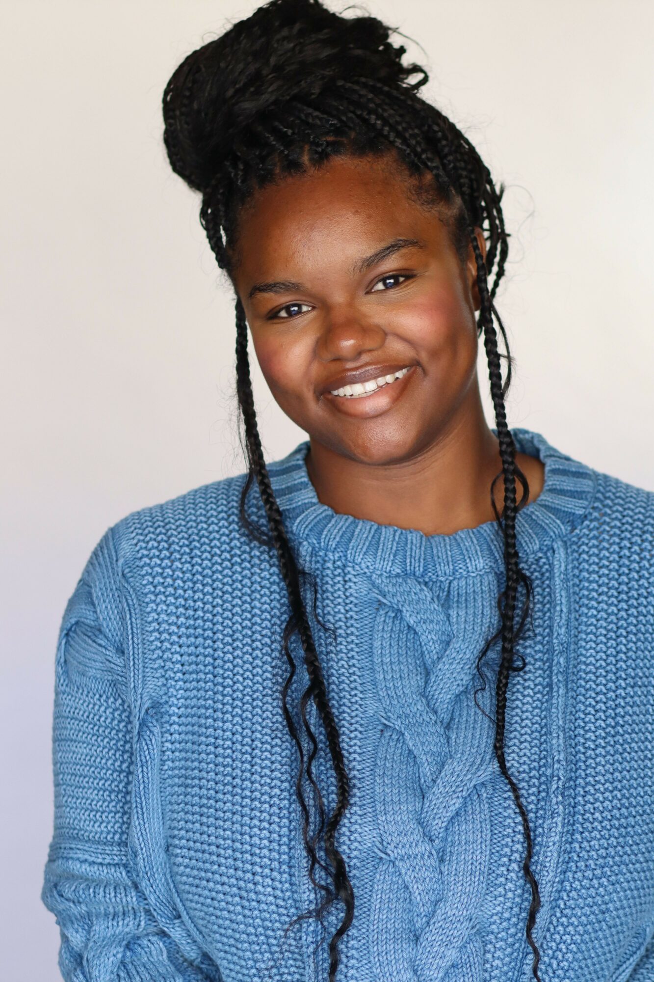 Smiling woman with braided hair wearing a blue sweater against a plain background.