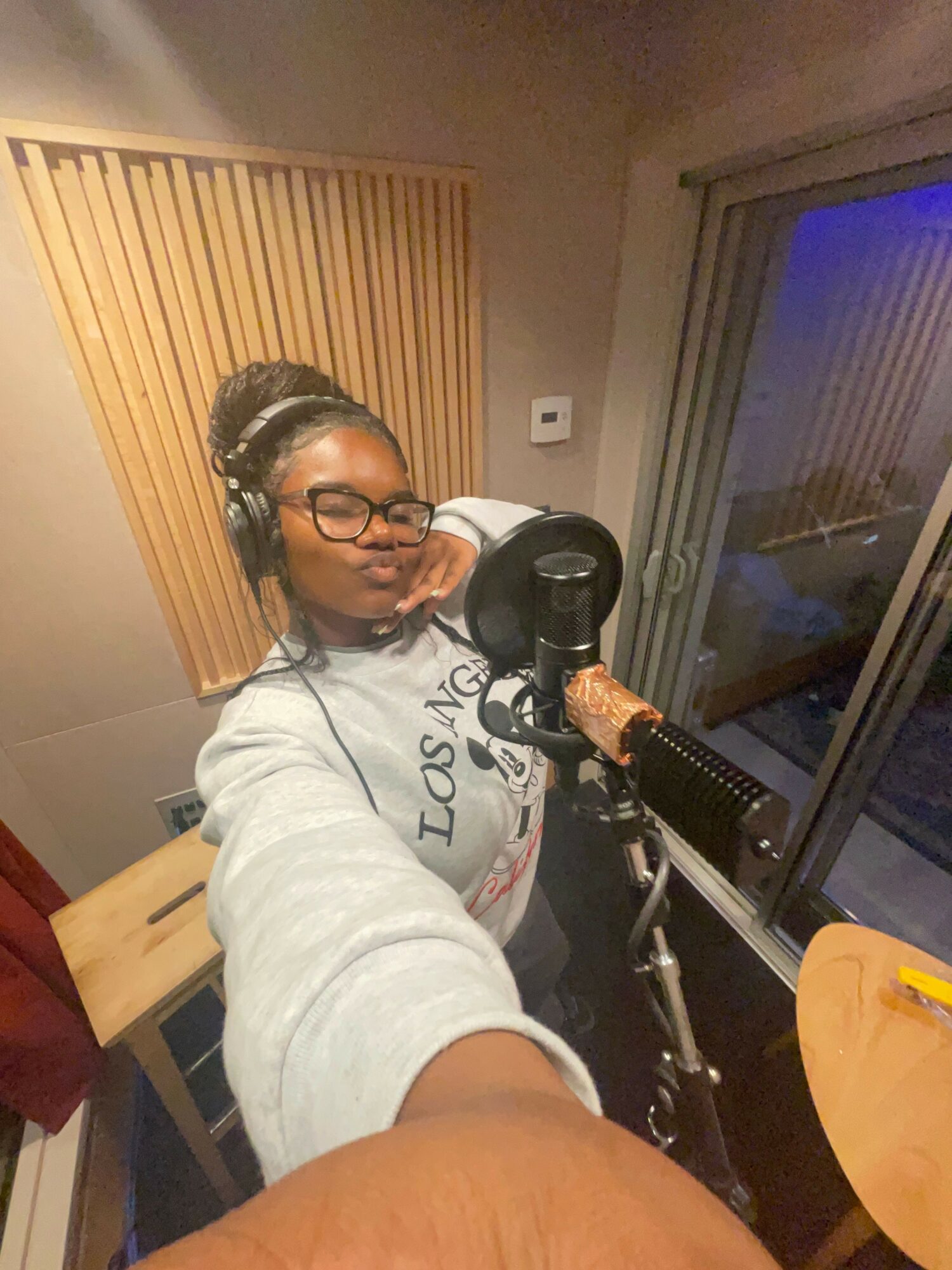 Woman with glasses and braided hair in a recording studio, taking a selfie with microphone and equipment.
