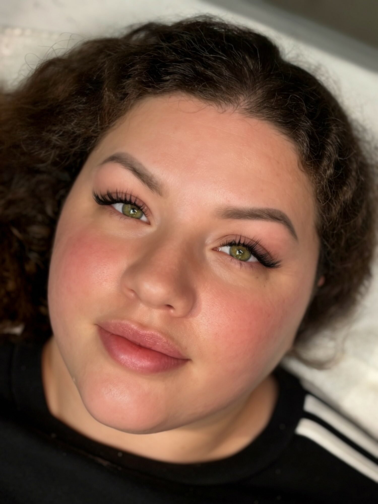 Close-up of a woman with curly hair, green eyes, and makeup, lying down and looking at the camera.