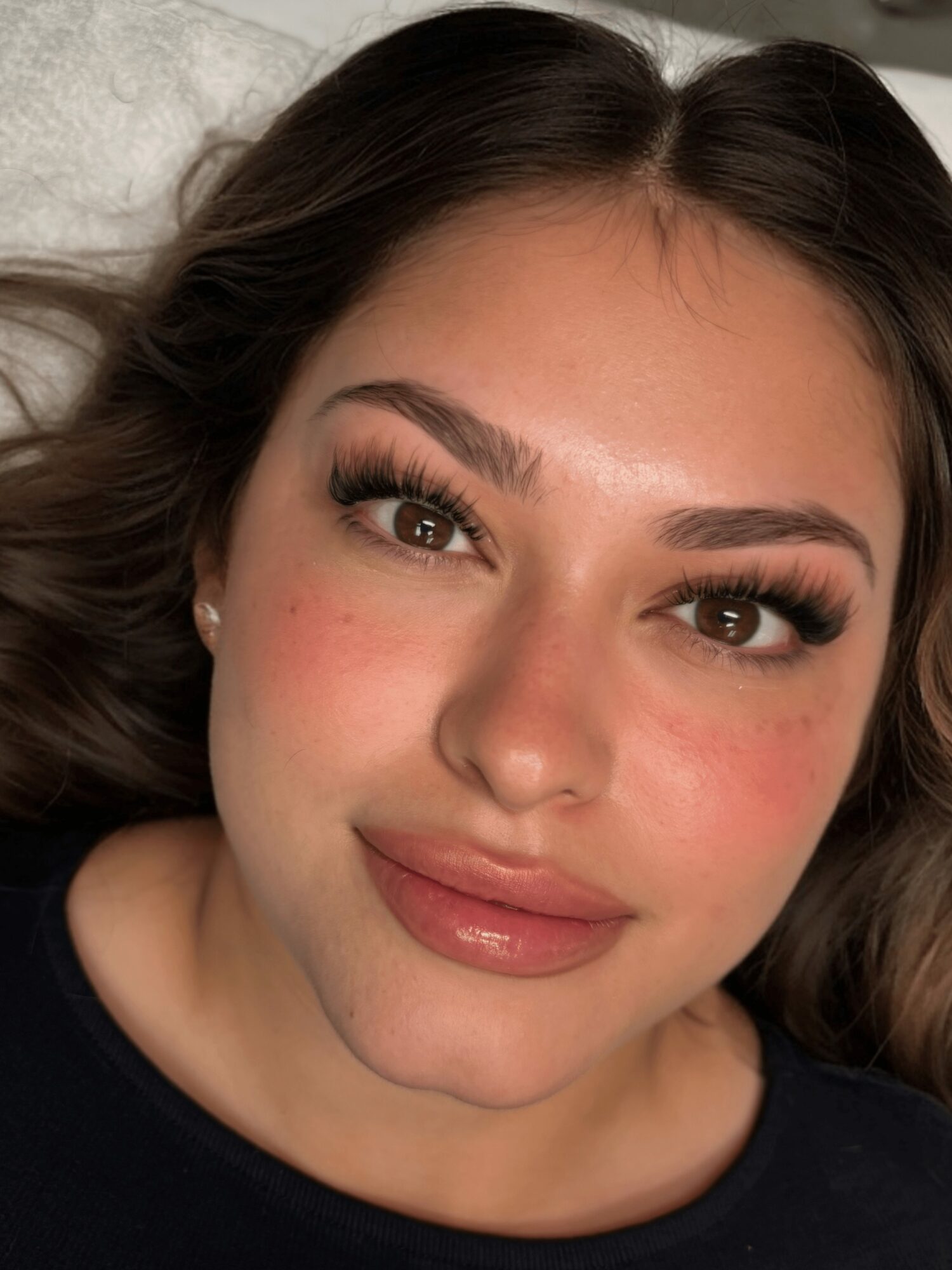 Close-up of a woman with long dark hair, makeup, and a slight smile, lying on a pillow.
