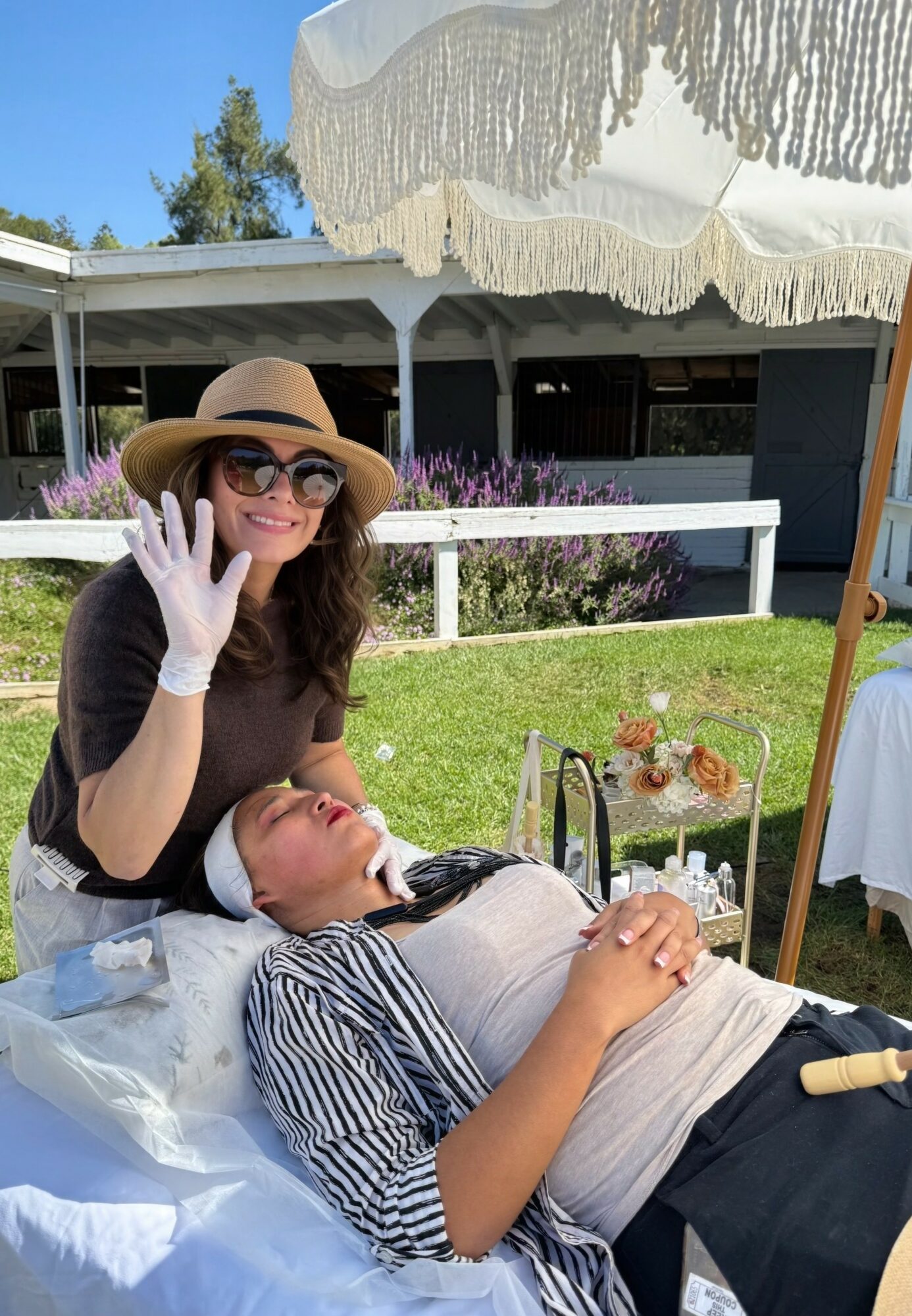 Woman wearing a hat, sunglasses, and white gloves smiling and waving, sitting beside a person lying on a lounge chair outdoors.