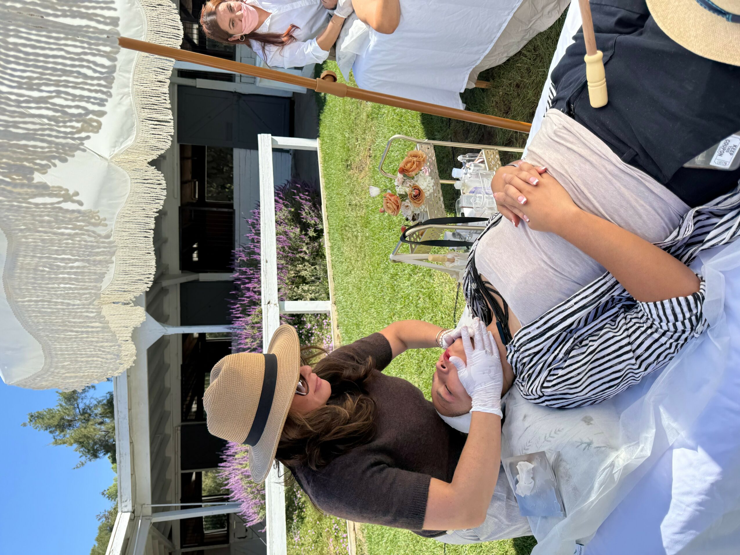 Two women sitting at a table outdoors, one with a hat, the other with glasses, engaged in conversation.
