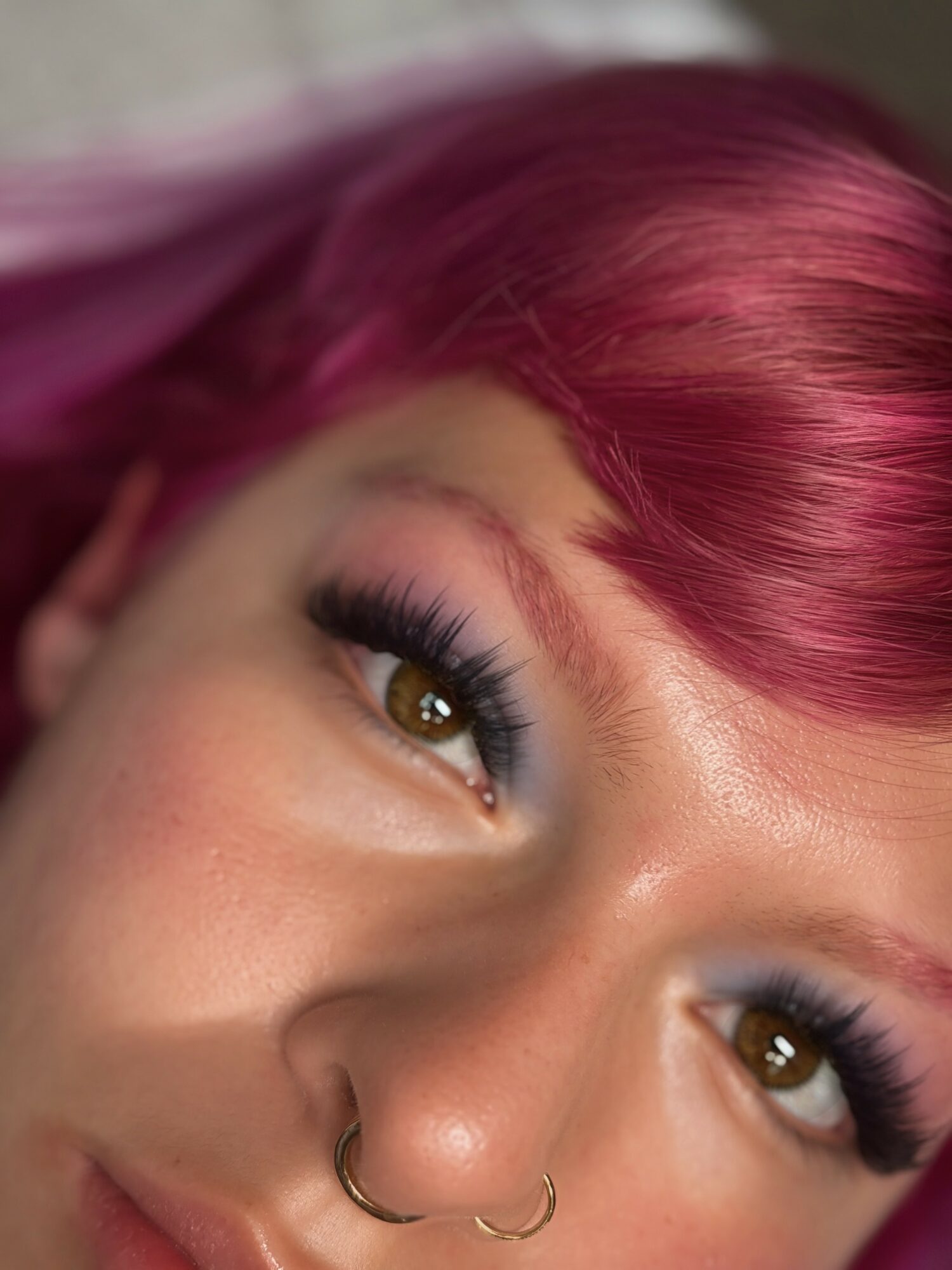 Close-up of a person's face with pink hair, brown eyes, and makeup including long eyelashes and colorful eyeshadow.