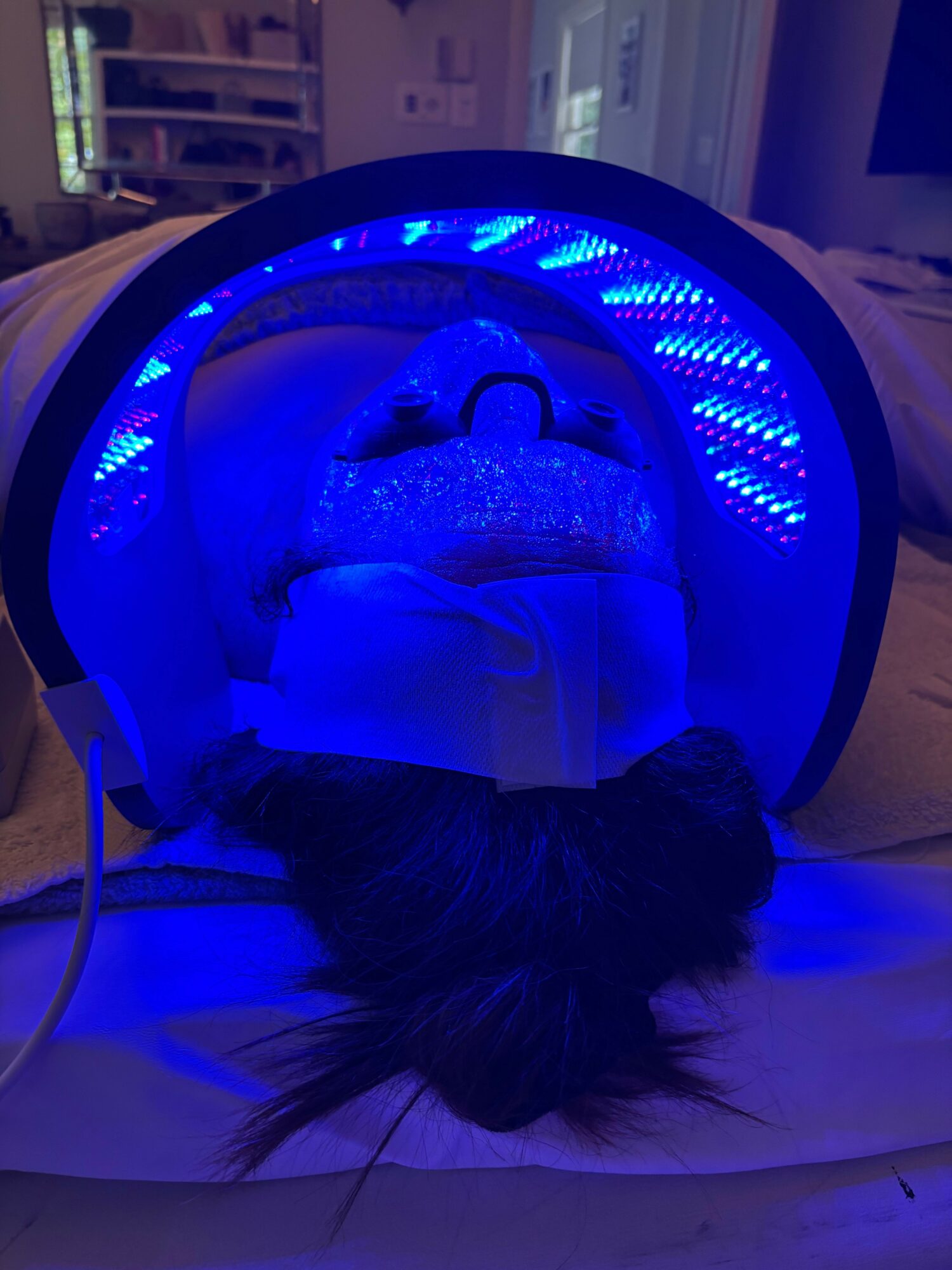 Person lying on bed with head under a blue LED light therapy device, hair visible at the bottom.