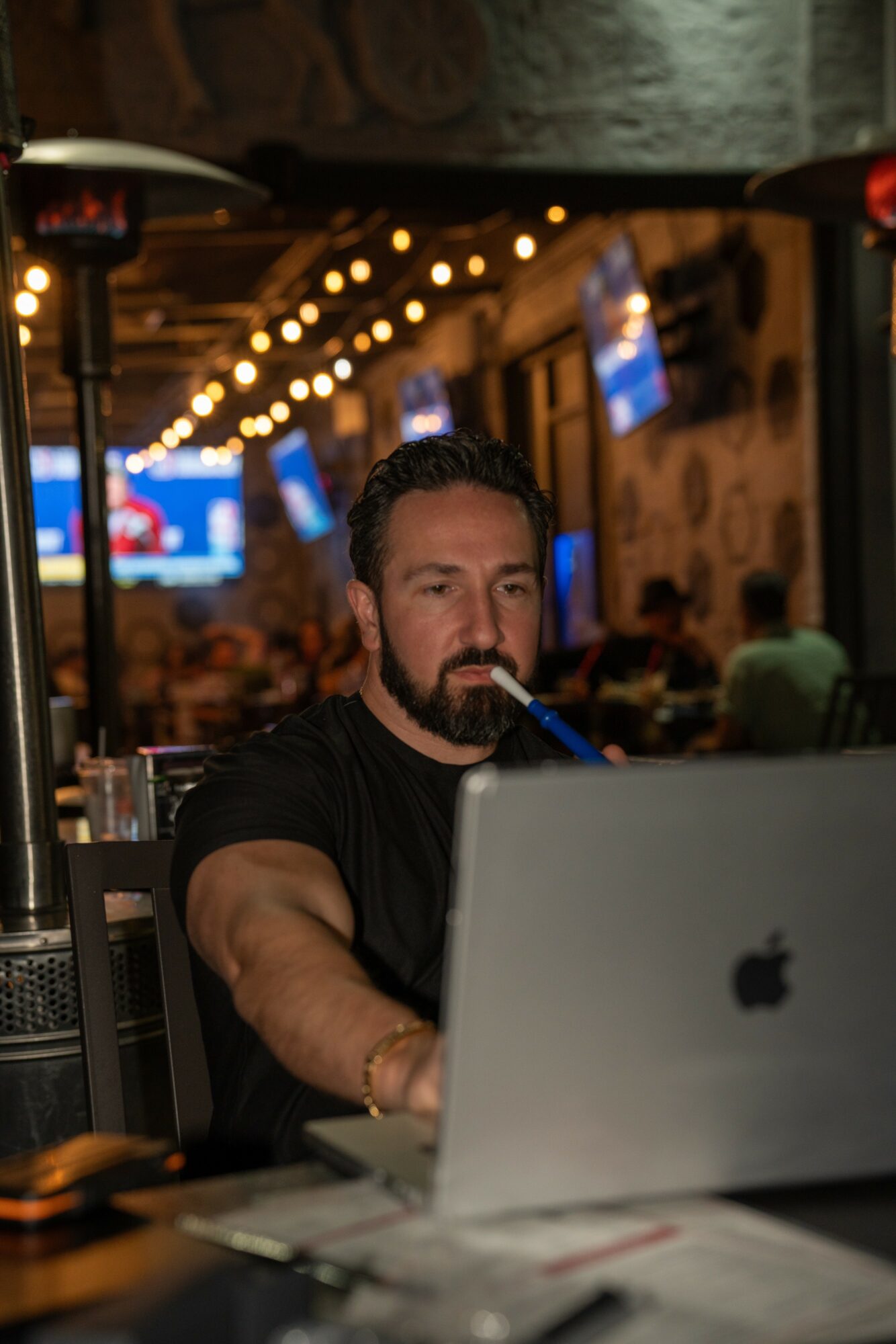Man with beard working on laptop in a dimly lit cafe with string lights and screens in background.