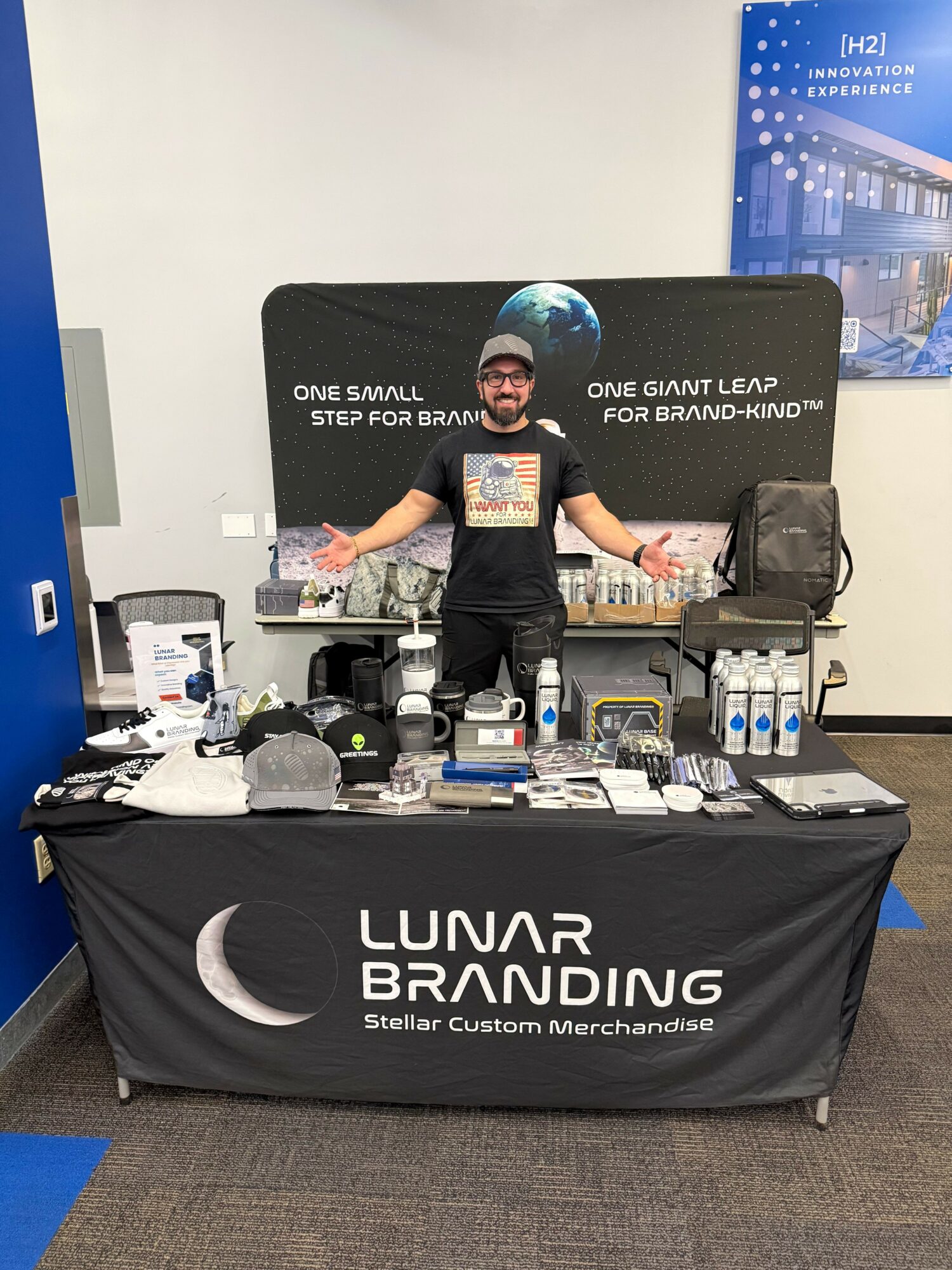 Person standing behind a table with branding merchandise, arms outstretched, at an indoor event booth.