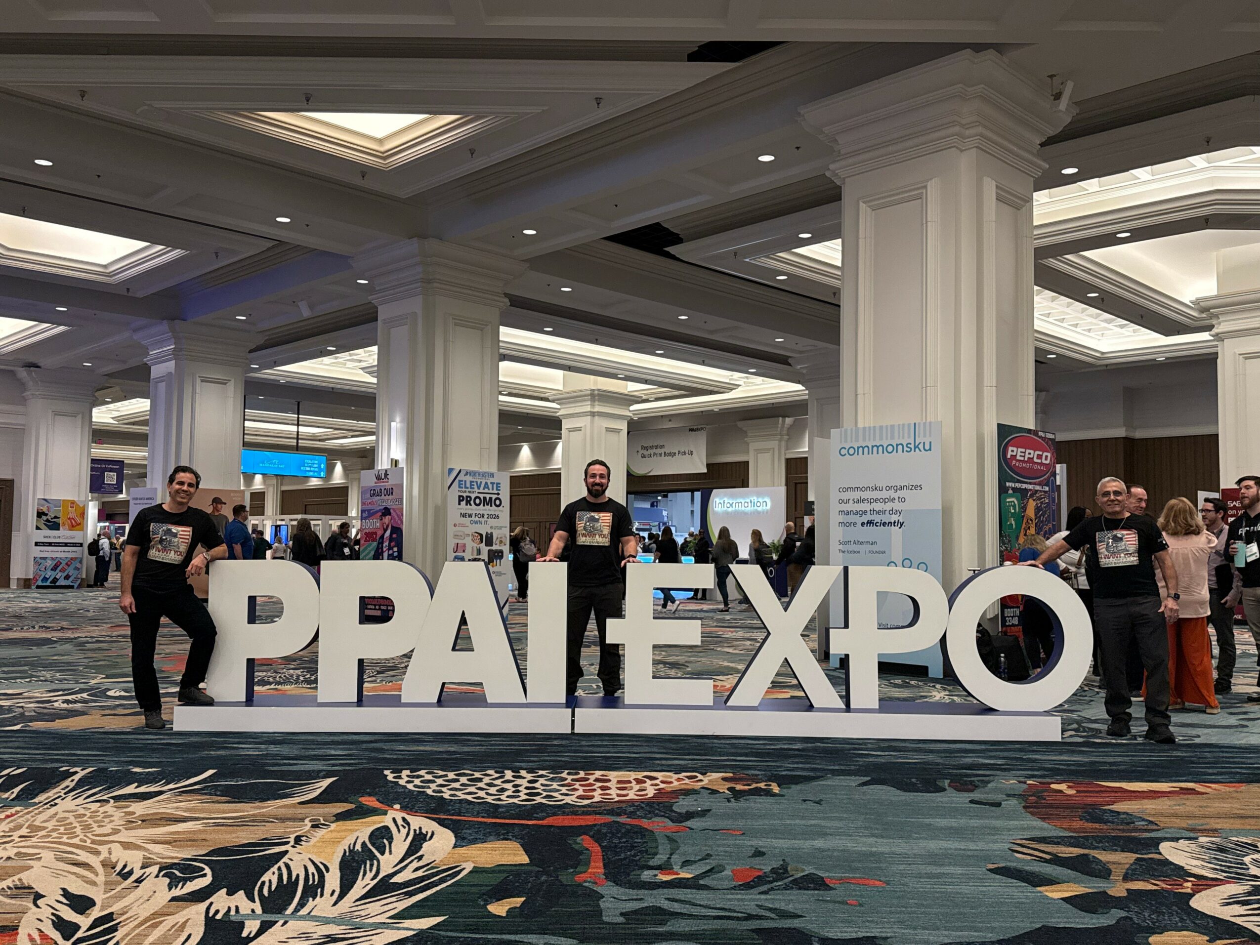 Large white letters spelling PPAI EXPO in a conference hall with people and booths in background.