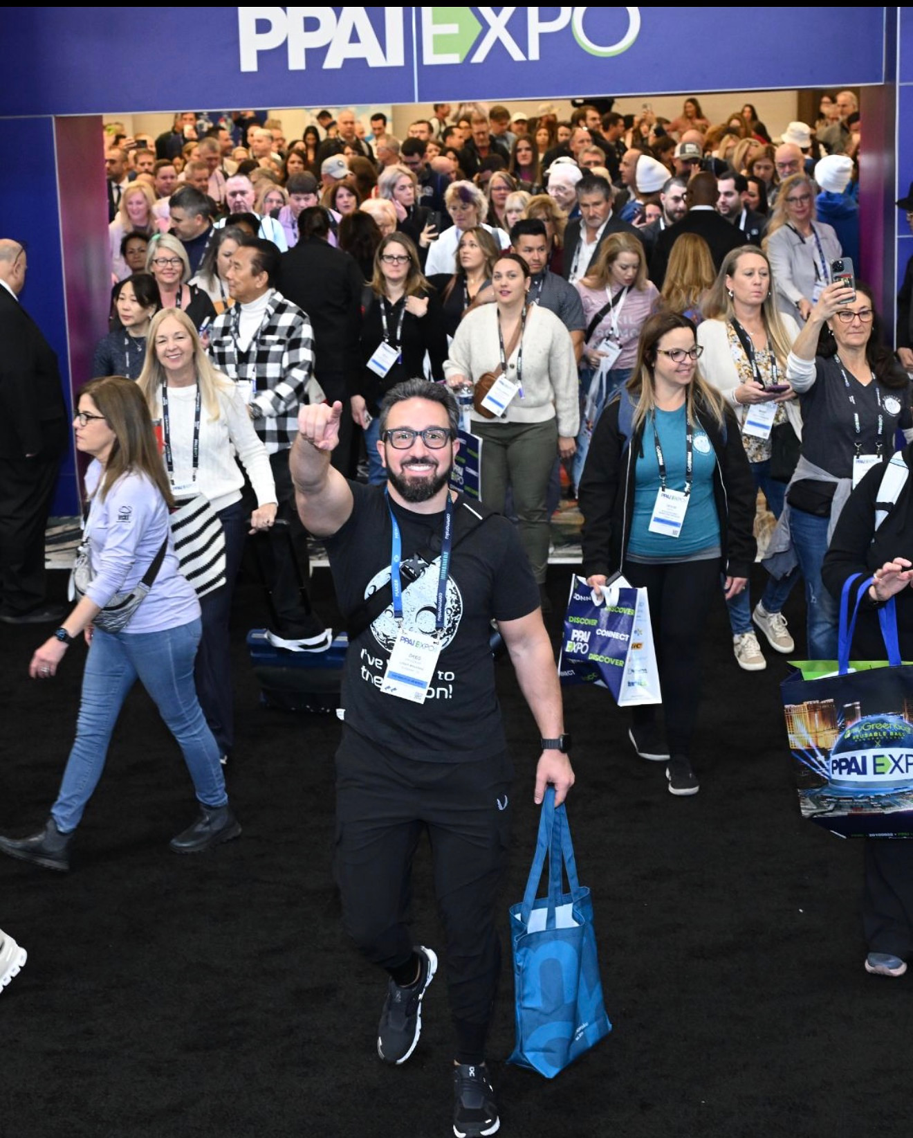 Man smiling and waving at a crowded conference event with many attendees in the background.