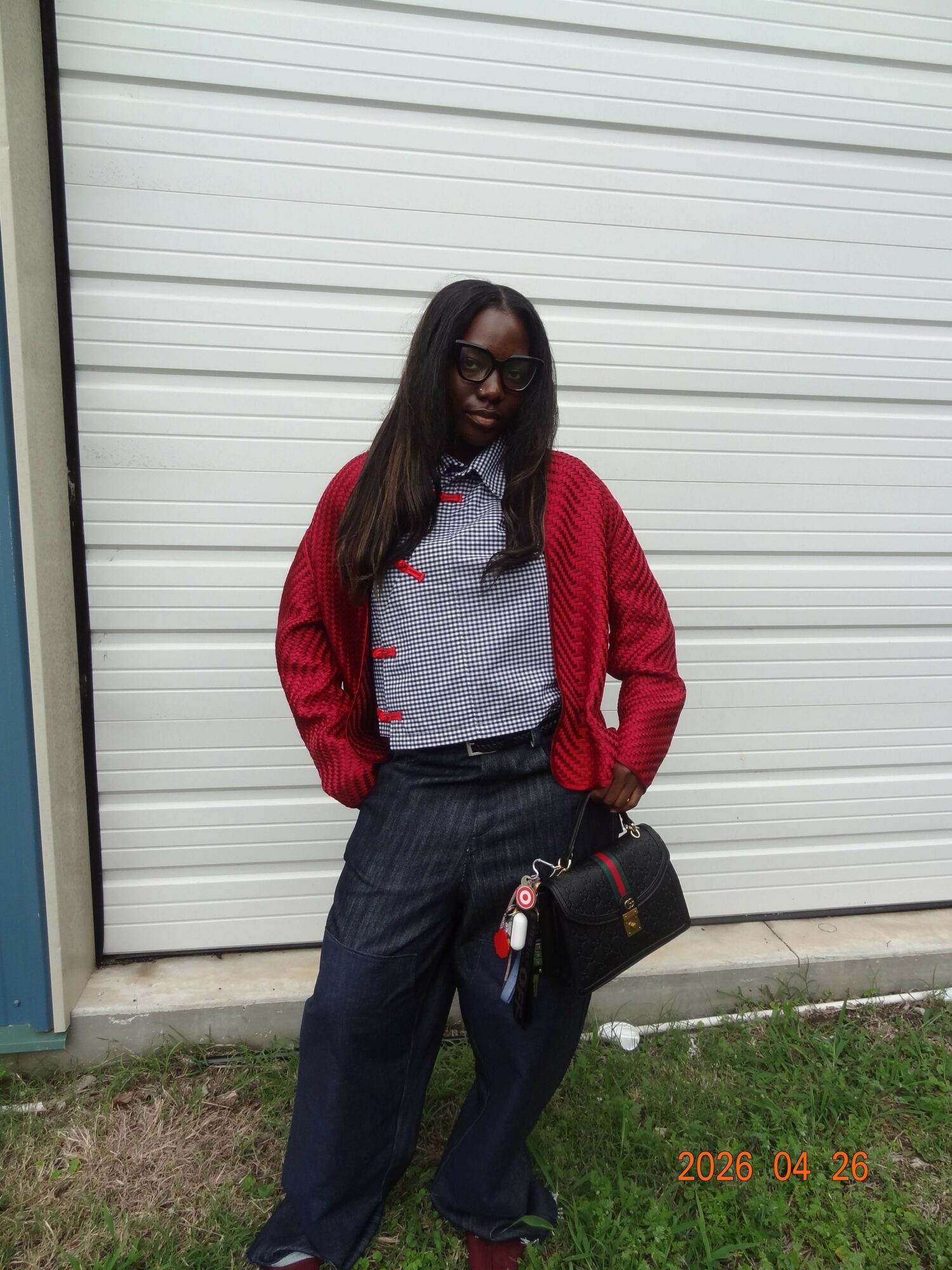 Person standing outdoors in front of a white garage door, wearing glasses, a red cardigan, gray shirt, and dark pants, holding a black bag.
