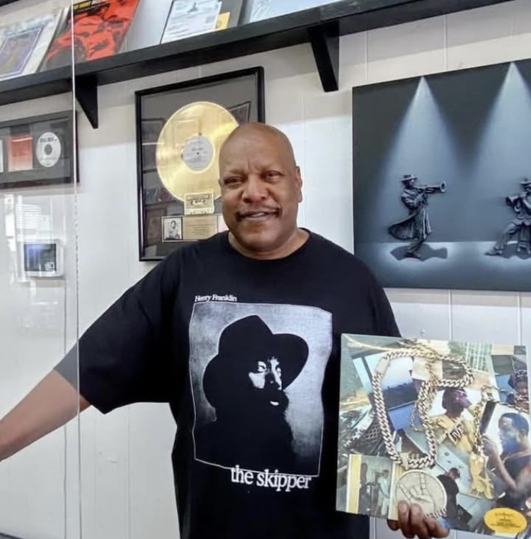 Man with a bald head smiling, wearing a black T-shirt with a silhouette and text, standing indoors near a display case.
