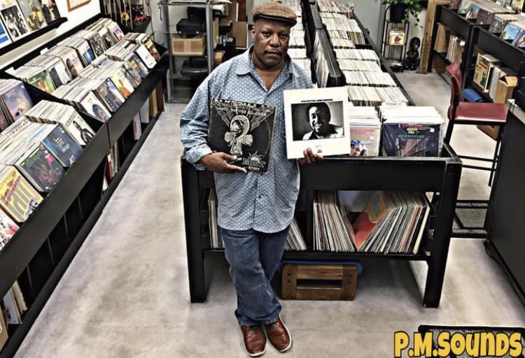 Man standing in record store holding vinyl records, surrounded by shelves of music albums.