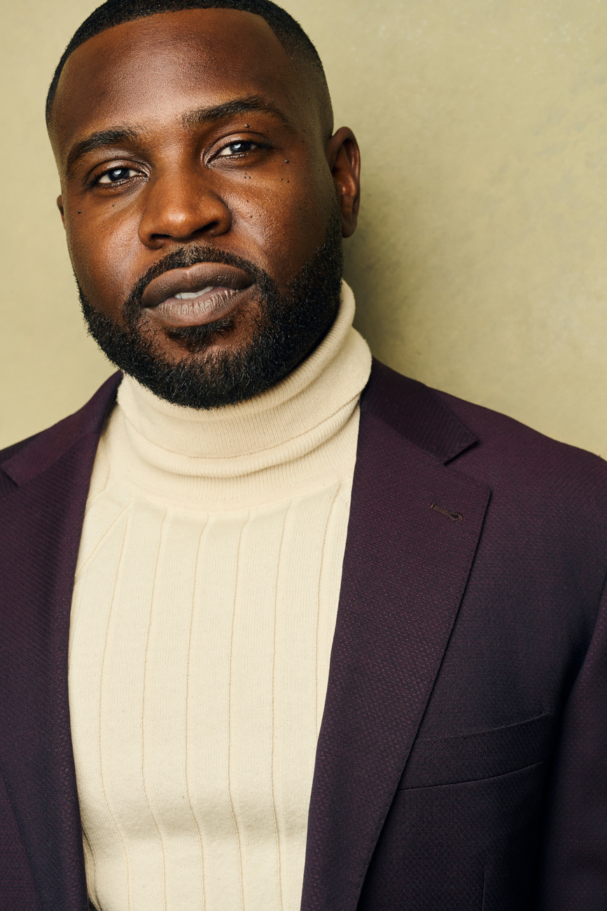 Man with dark skin, beard, wearing a white turtleneck and dark blazer, looking at the camera.