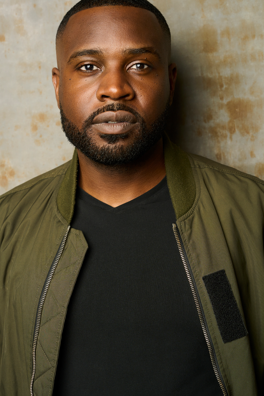 Man with short hair and beard wearing a green jacket and black shirt, standing against a textured wall.