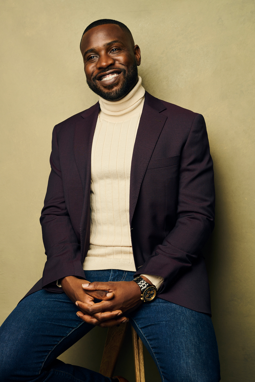 Smiling man with a beard in a dark blazer and cream turtleneck, sitting against a plain background.