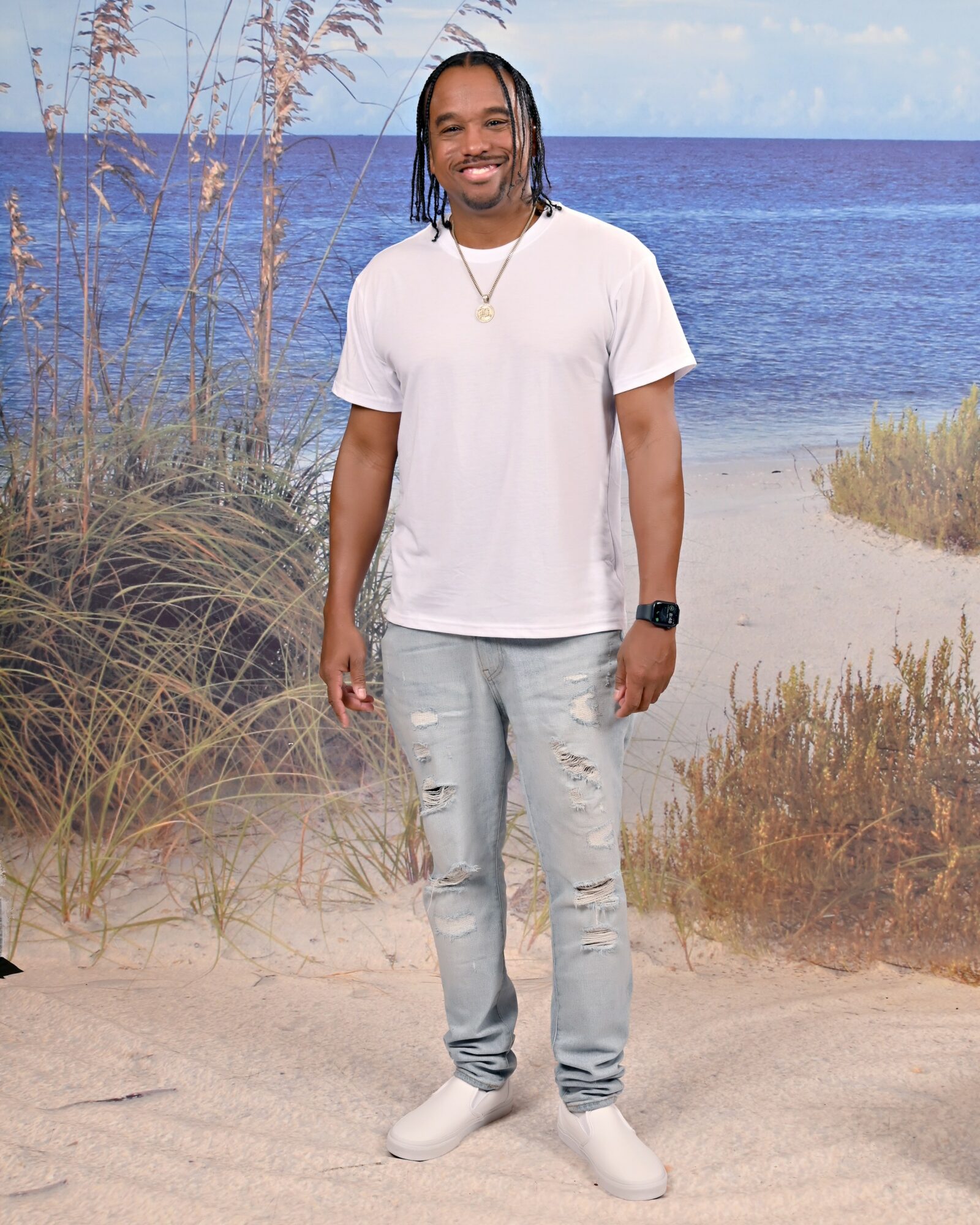 Young man standing on sandy beach with ocean and sky in background, smiling, wearing white t-shirt and light jeans.