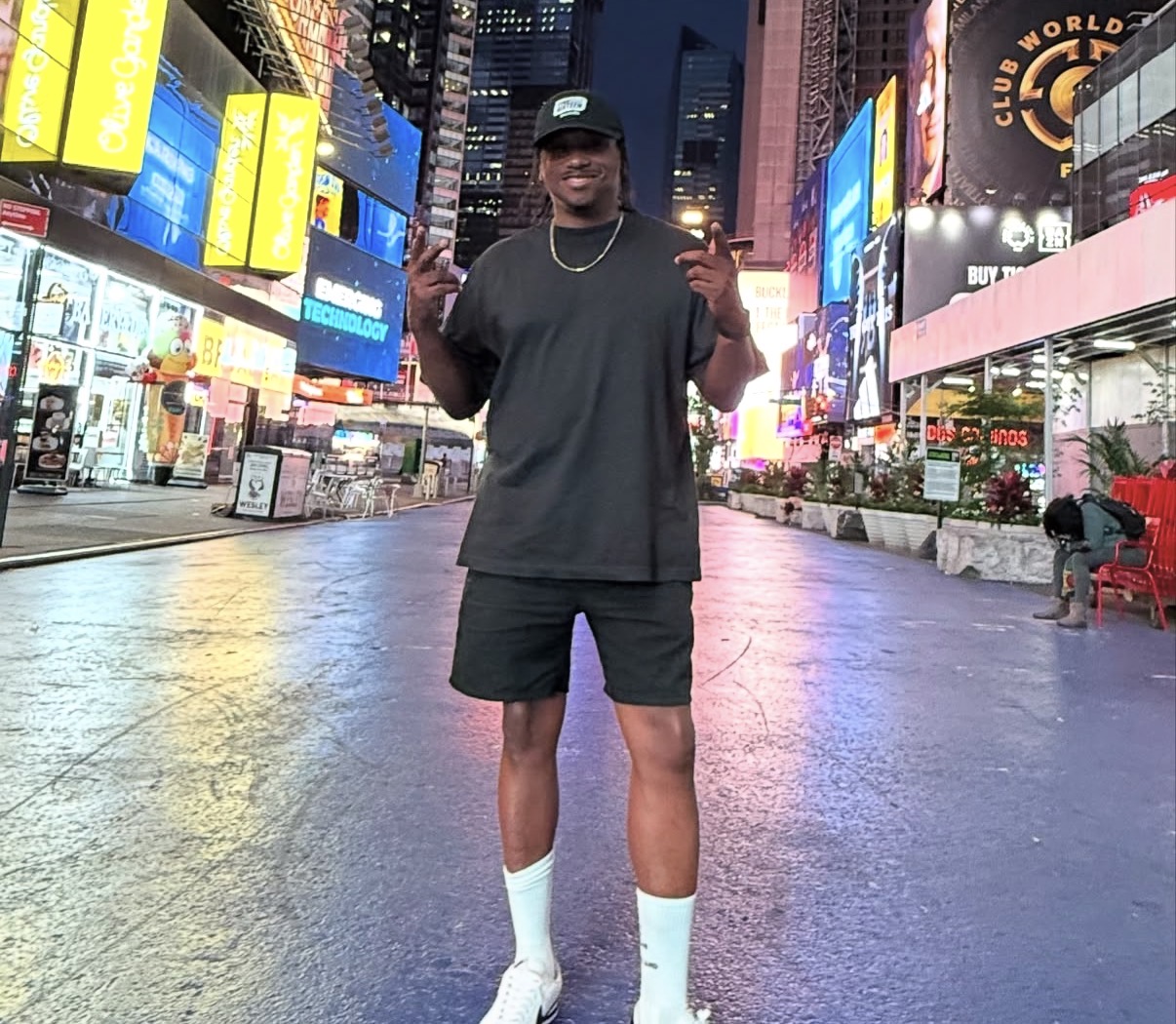Young man standing in Times Square at night, smiling, wearing a black t-shirt, shorts, white socks, and a cap.