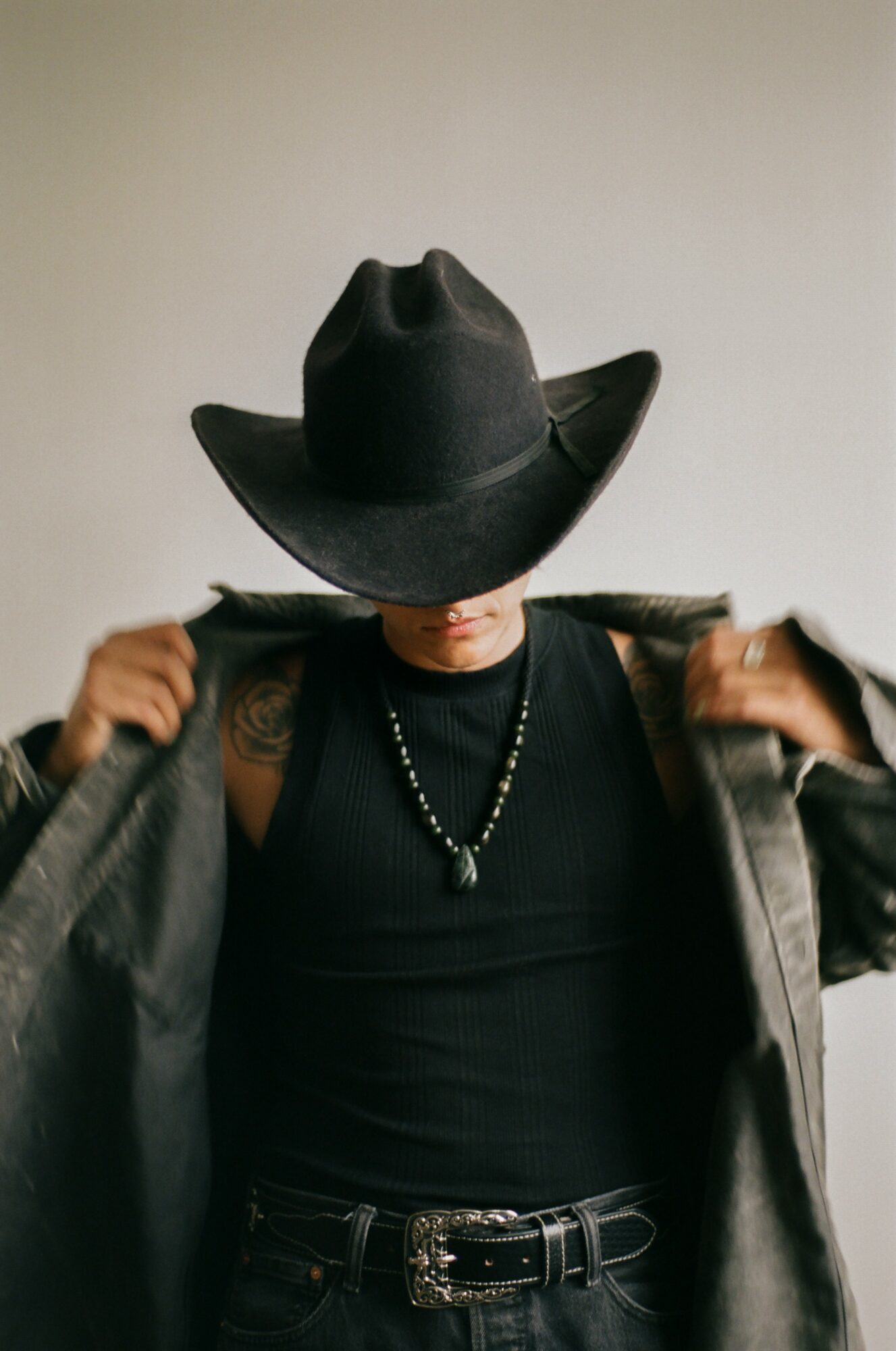 Person wearing a wide-brimmed hat, black shirt, and necklace, adjusting a jacket over shoulders.