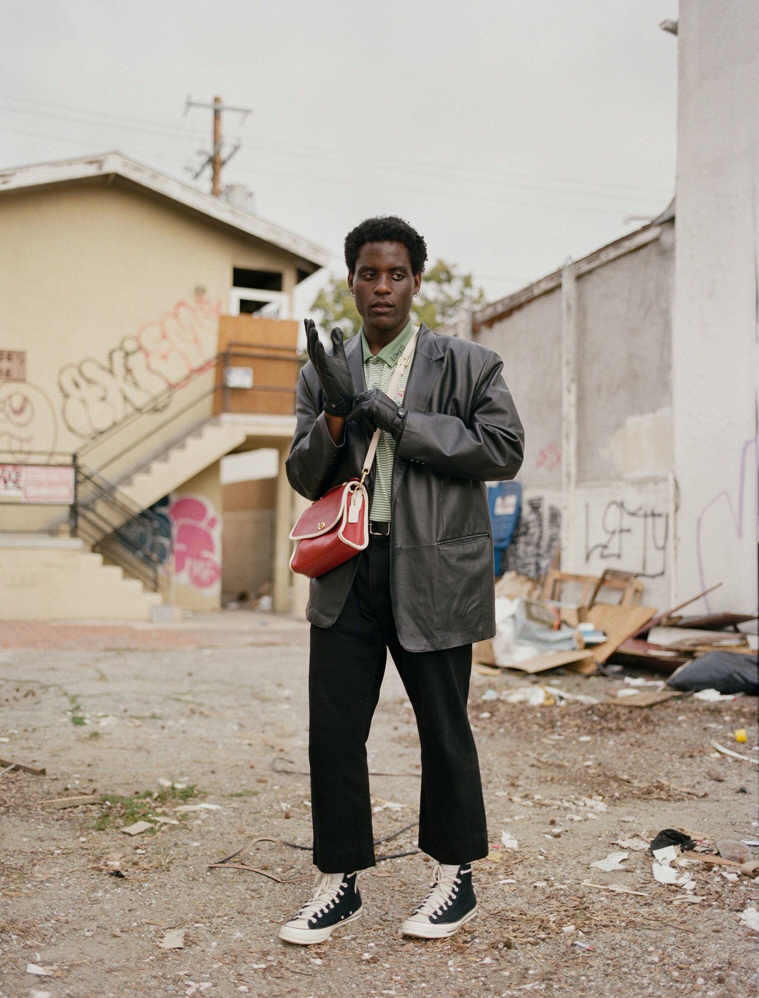 Young person standing outdoors in front of graffiti-covered wall, wearing a black jacket, black pants, and sneakers, holding gloves.