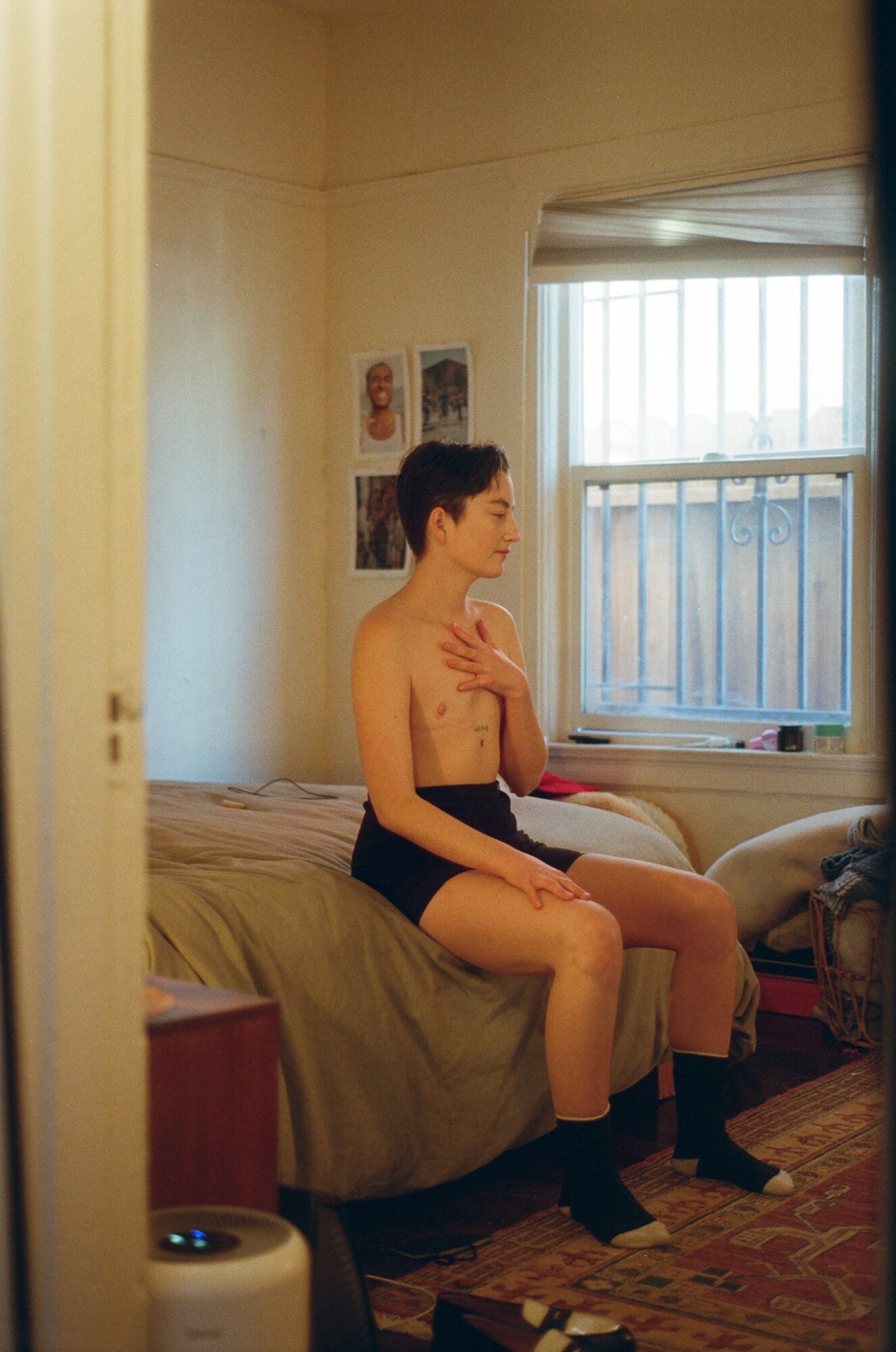 Young person sitting on bed with hand on chest, in a room with window and wall decorations.