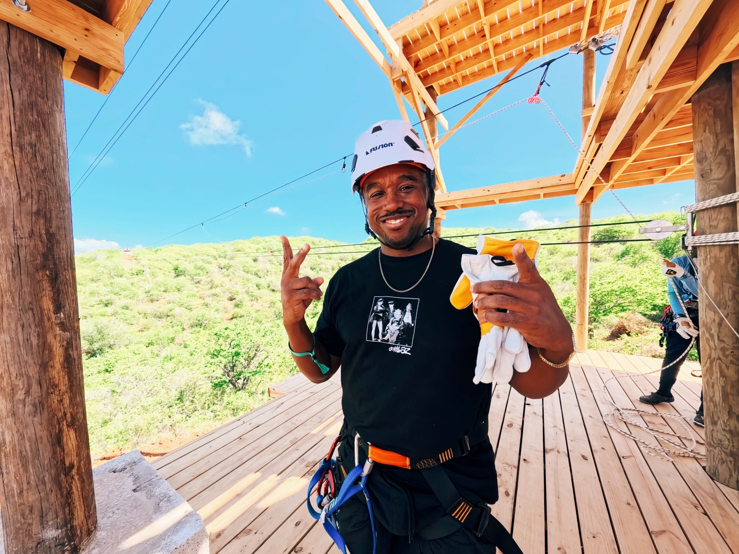 Smiling man wearing a helmet and harness, holding a yellow object, on a wooden platform outdoors with blue sky and greenery.