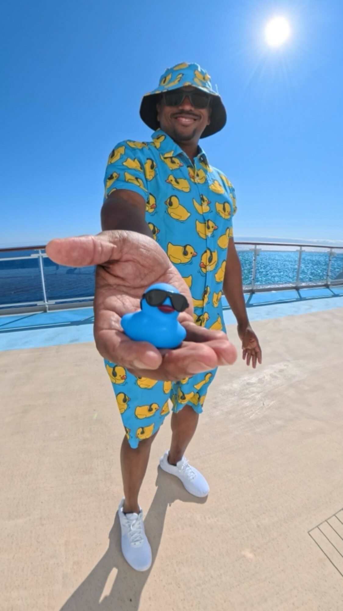 Person in a blue and yellow patterned outfit holding a blue rubber duck on a cruise ship deck under a sunny sky.