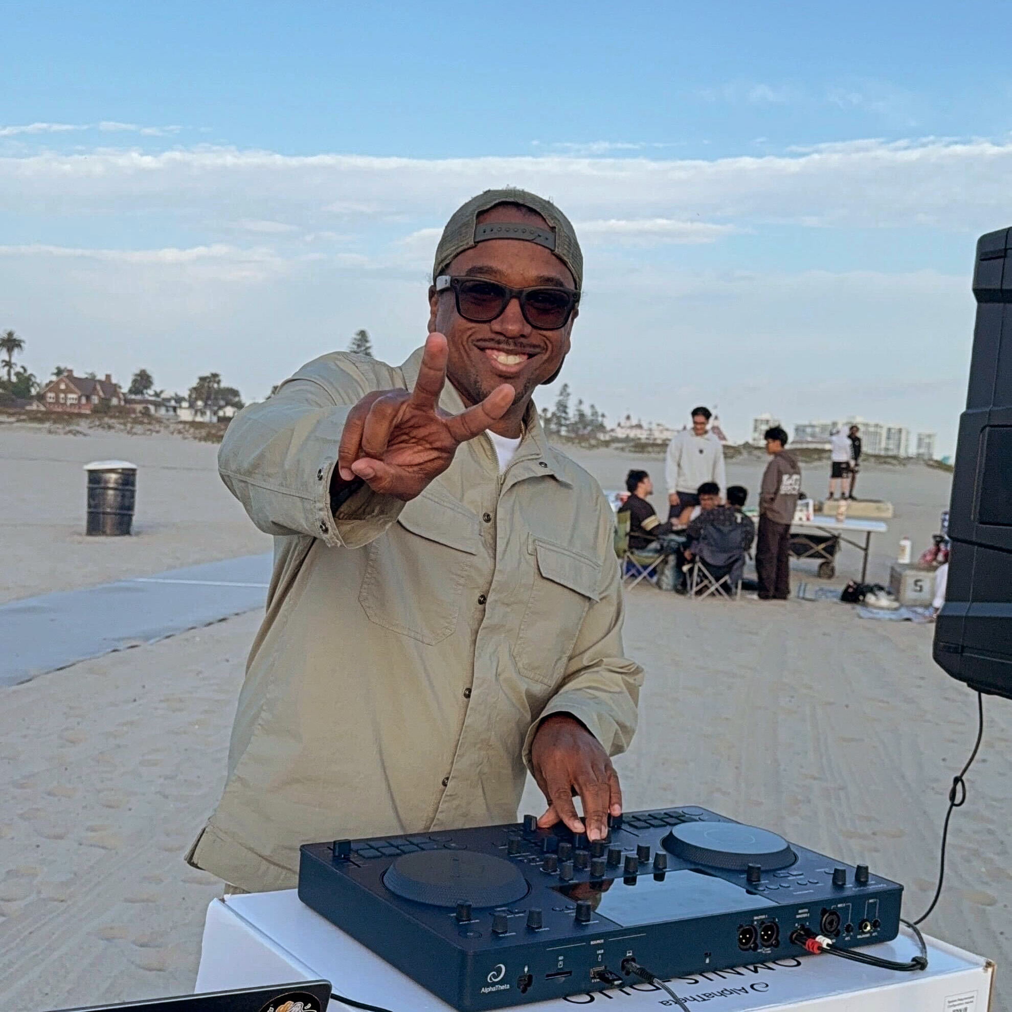 Person wearing sunglasses and a cap, smiling, standing outdoors with a DJ equipment, people in the background.