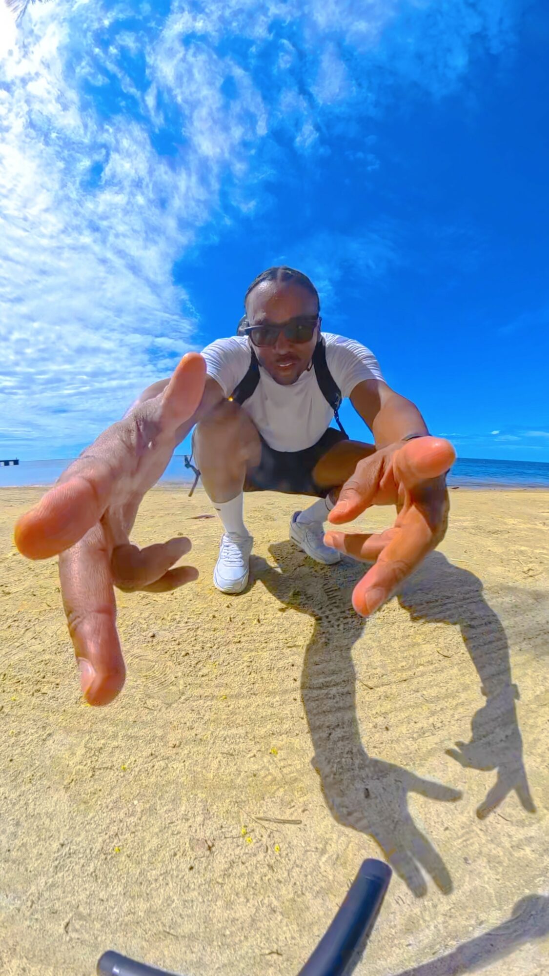Person crouching on sandy beach, reaching towards camera with hands, blue sky with clouds overhead, ocean in background.