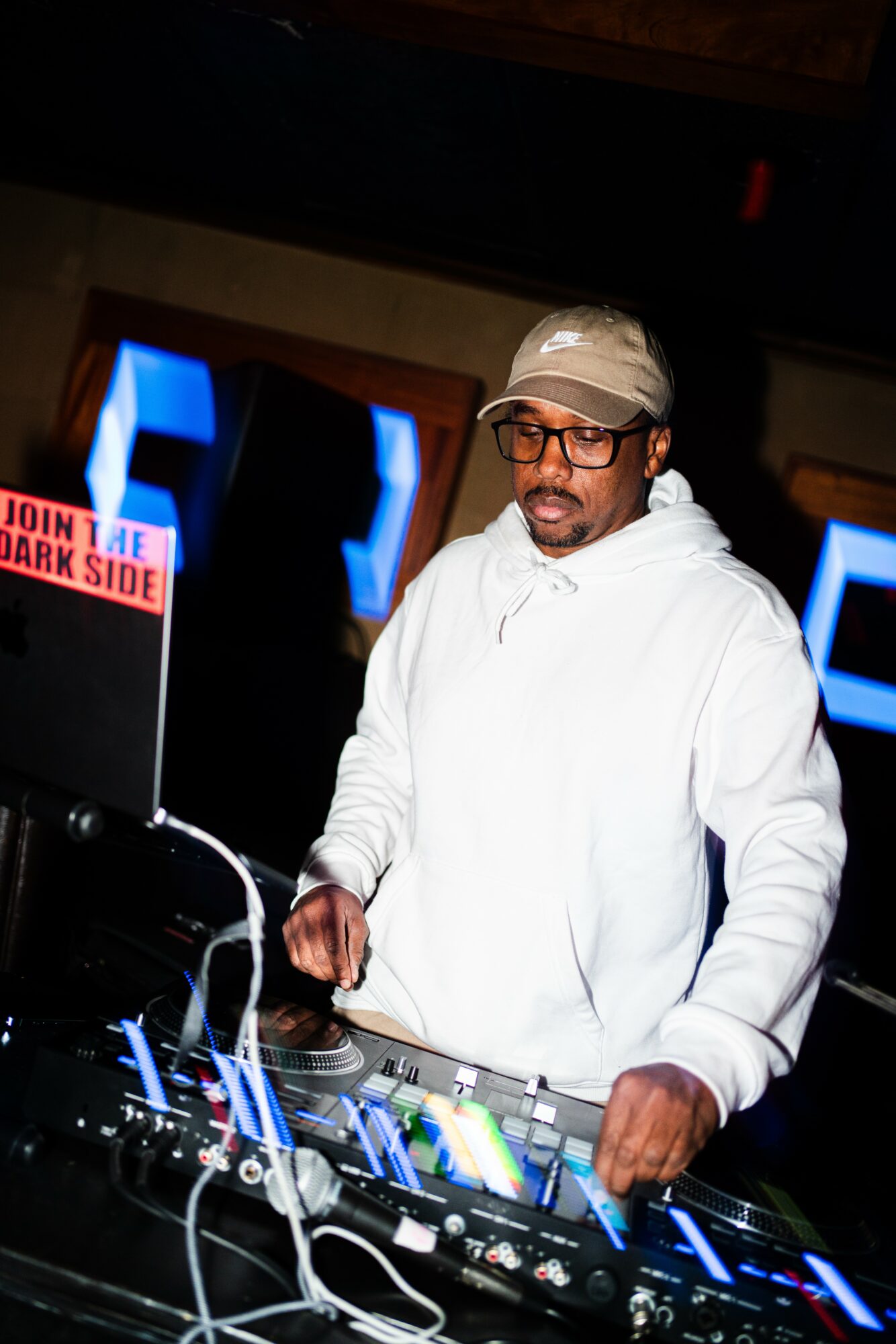 Man in white hoodie and cap operating DJ equipment with colorful lights, dark background, and a sign reading 'Join the Dark Side'.