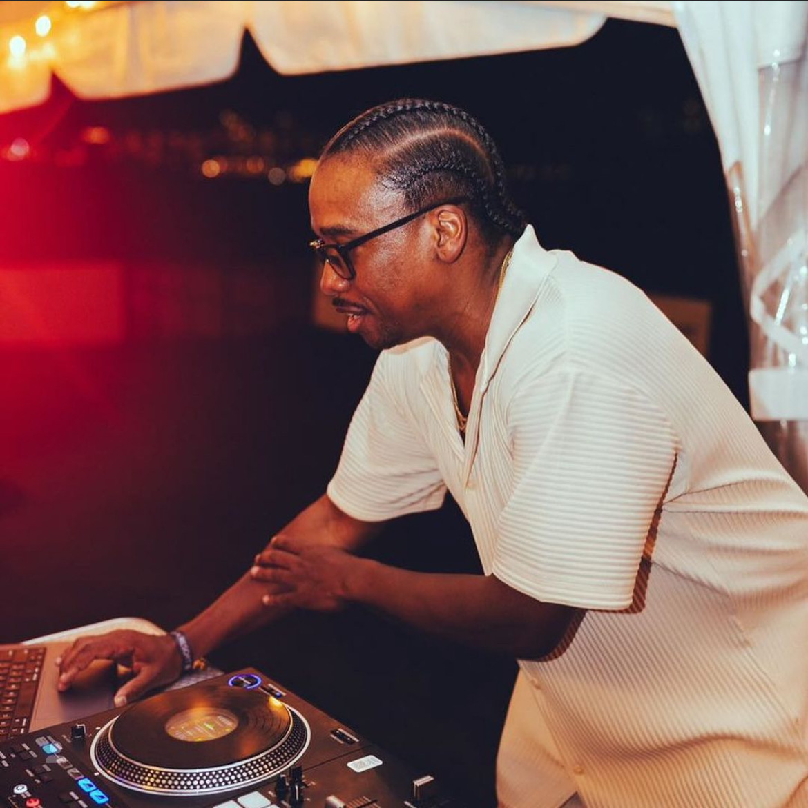 Person with glasses and braided hair working as a DJ at a table with equipment, in a dimly lit setting.