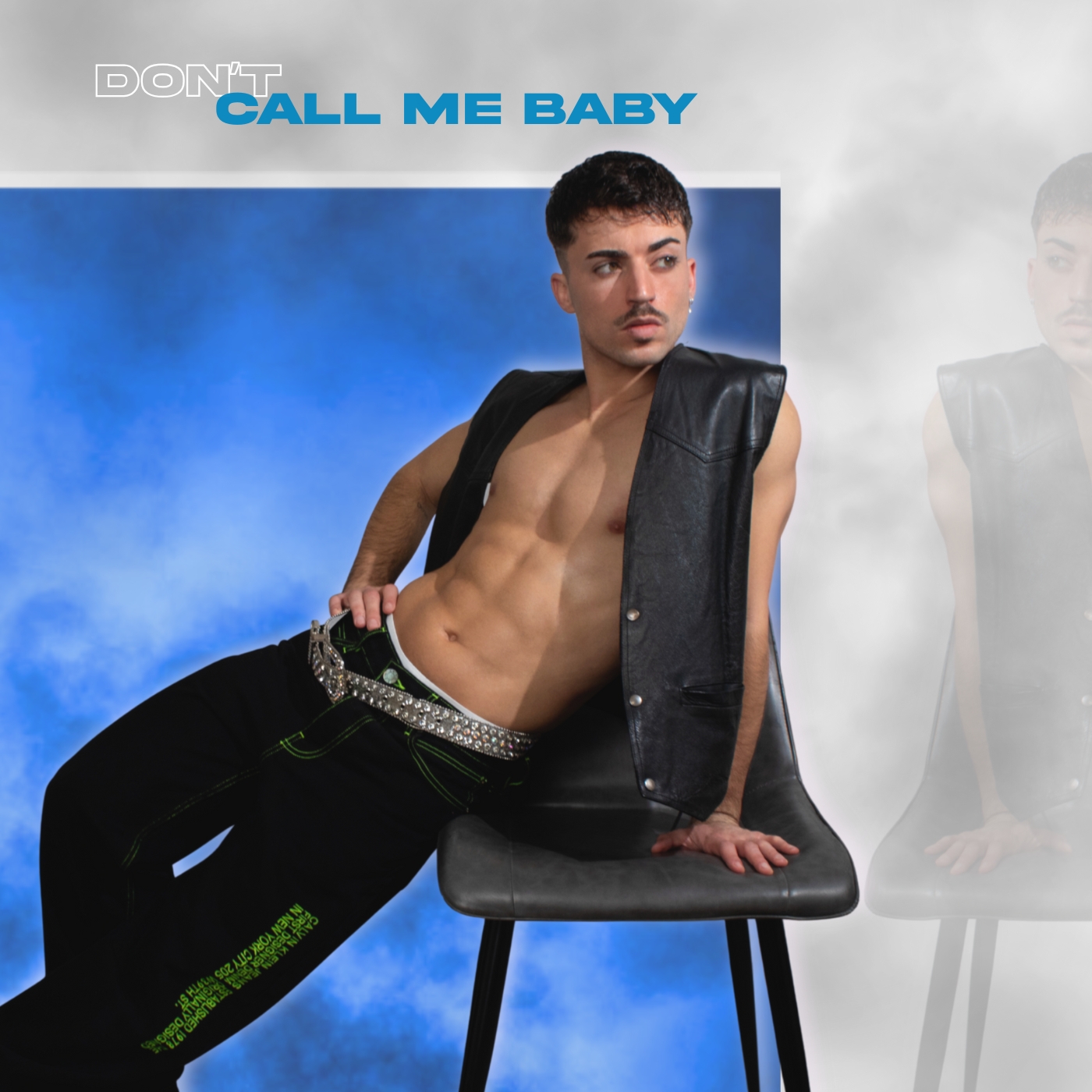 Young man with short dark hair and mustache, shirtless, wearing a black vest and pants, sitting on a black chair.