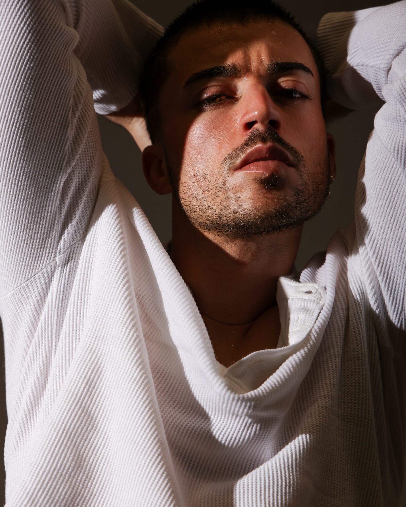 Man with short dark hair and stubble, wearing a white shirt, looking directly at camera with serious expression.