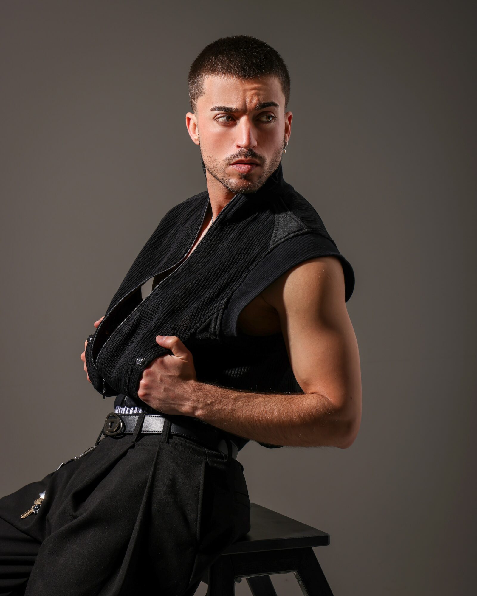 Young man with short hair and beard, wearing a black sleeveless jacket, sitting on a stool against a gray background.