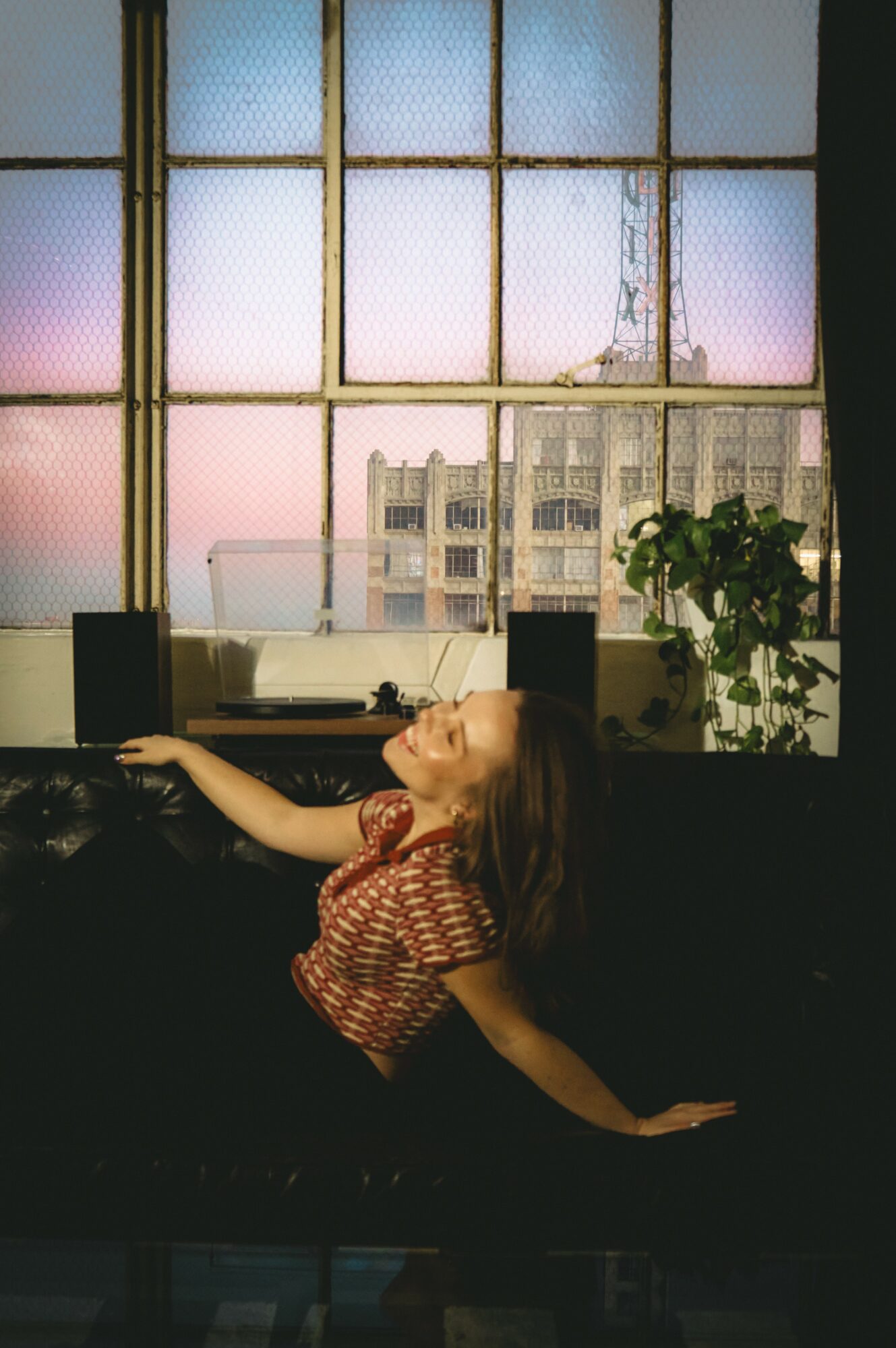Woman with long hair reclining on a dark surface near a large window with a cityscape view.
