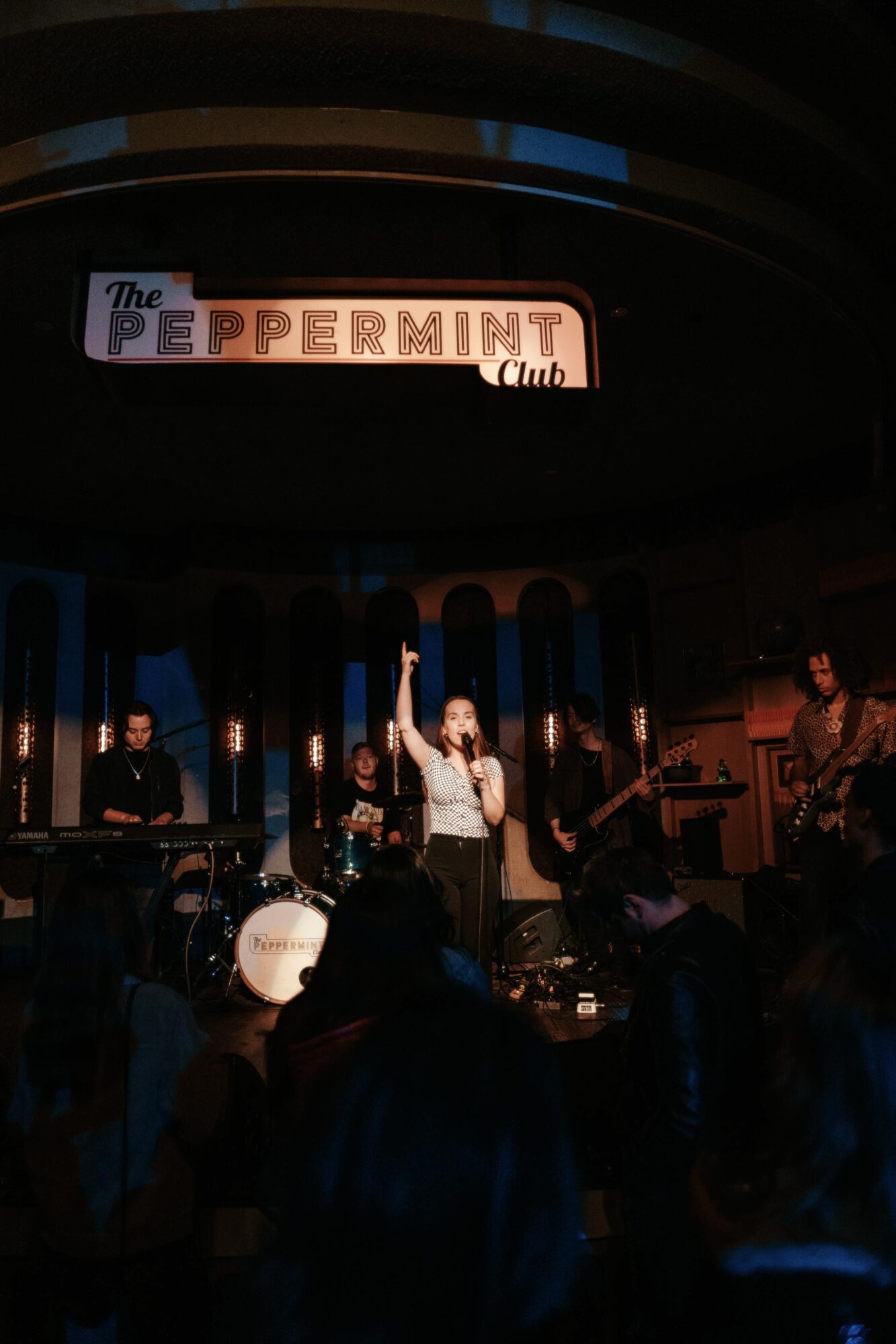 Band performs on stage with a singer raising a hand, audience watching, sign above reads 'The Peppermint Club'.
