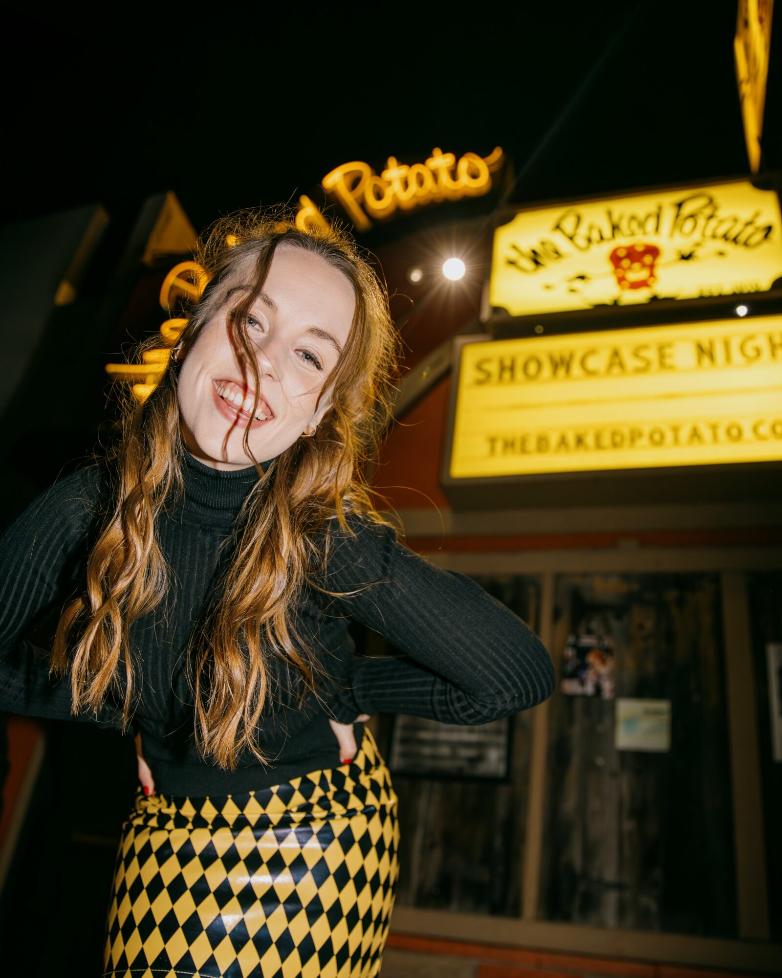 Young woman with long hair smiling and posing outdoors at night, illuminated by bright lights and signs.
