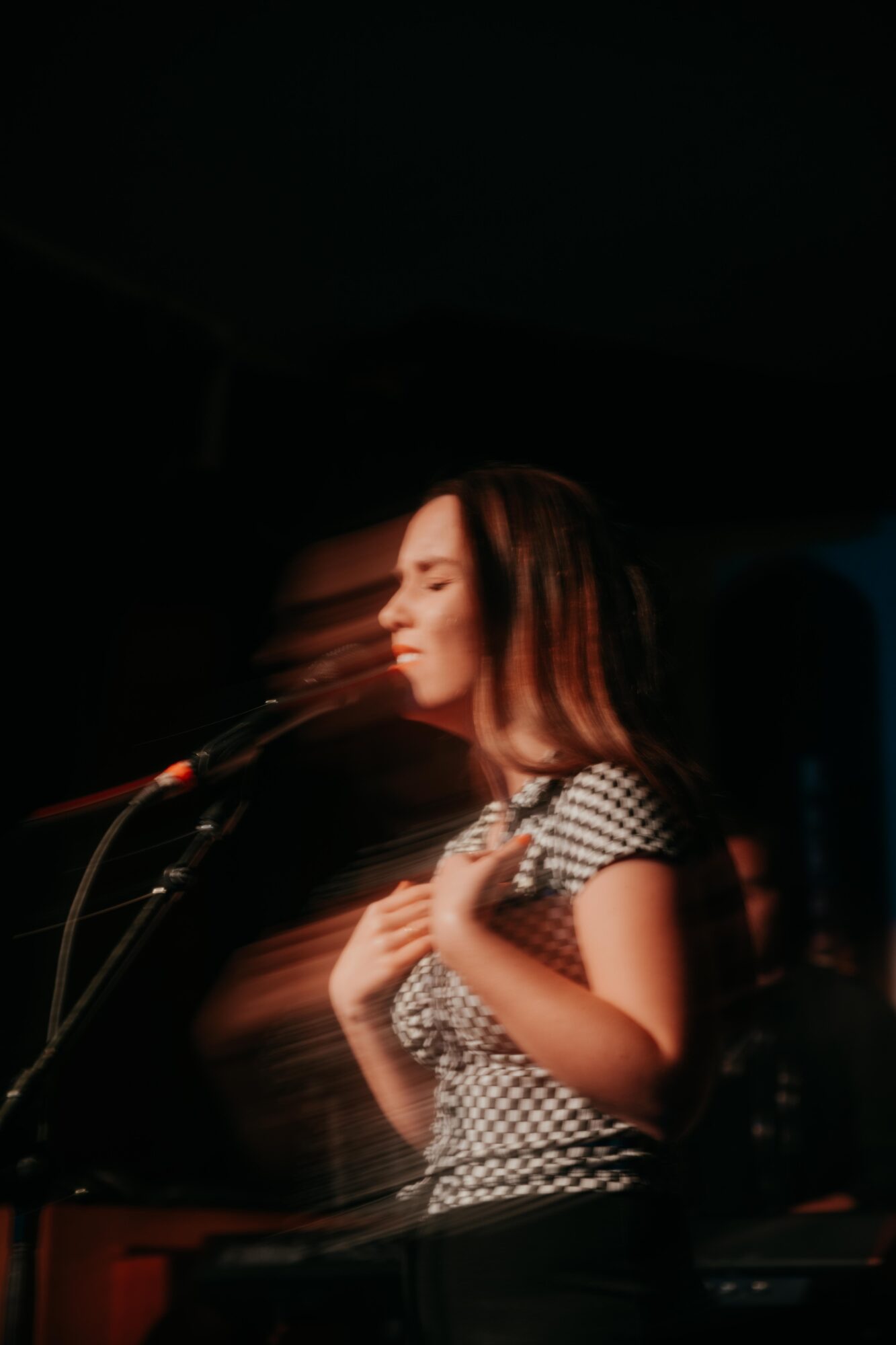 Young woman with long hair singing into a microphone on stage, wearing a checkered top, with motion blur effect.