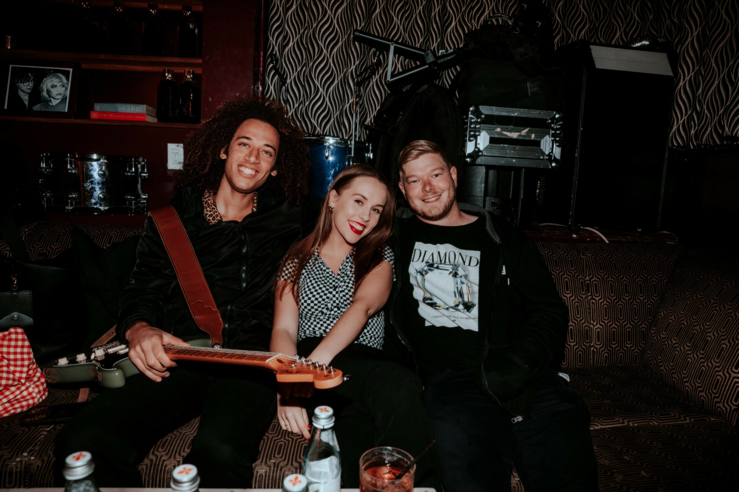 Three people sitting together on a couch, smiling, in a dimly lit room with musical equipment in the background.