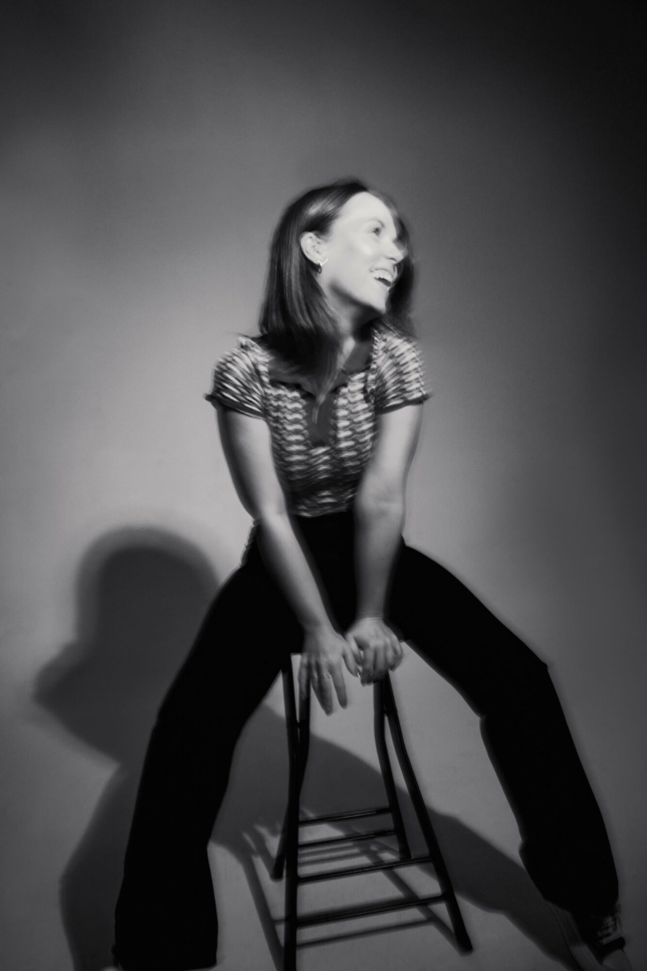 Woman sitting on a stool, leaning forward with hands on knees, in a black and white photo.