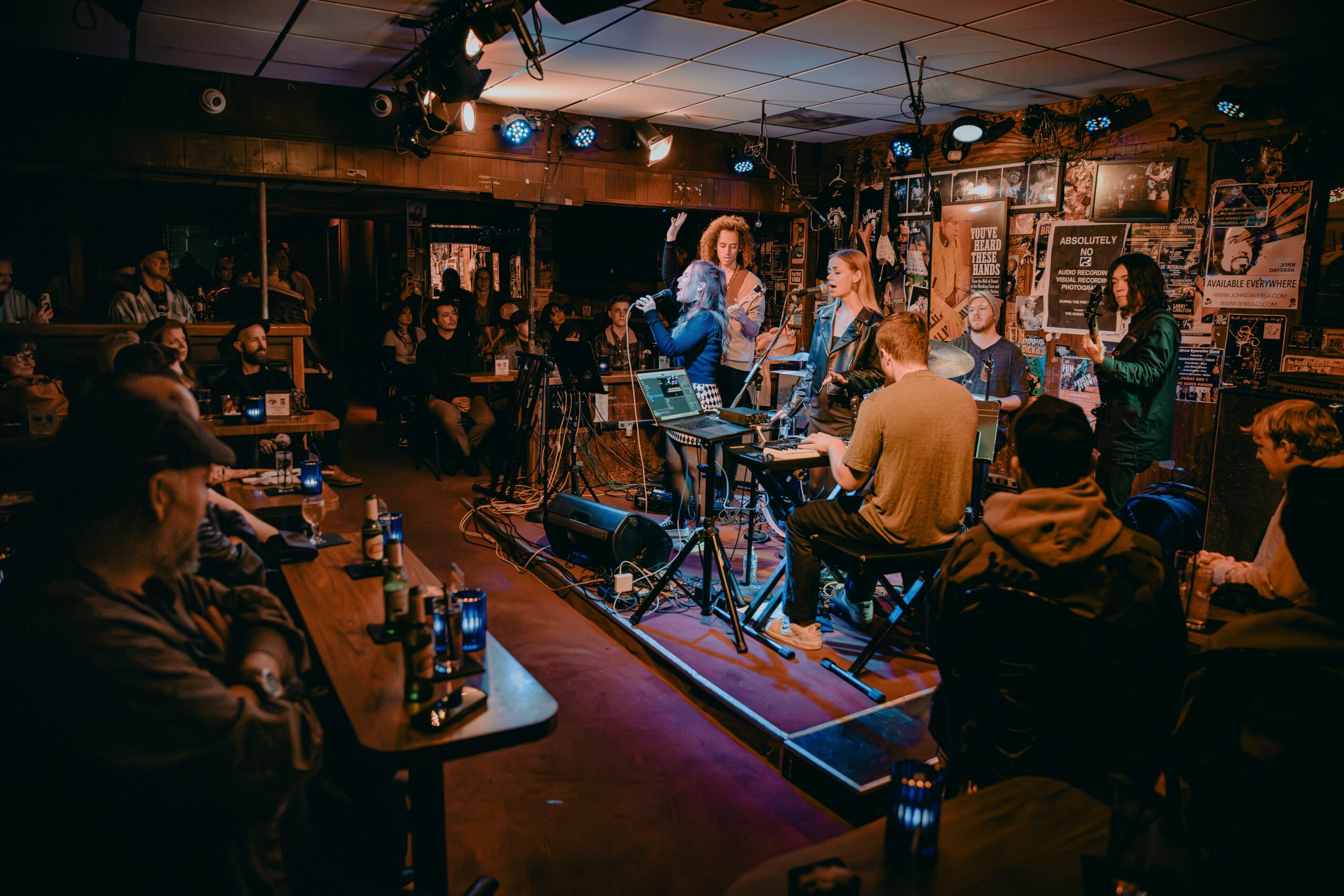 Live music performance with a singer and band on stage in a dimly lit venue, audience seated at tables, some with drinks.