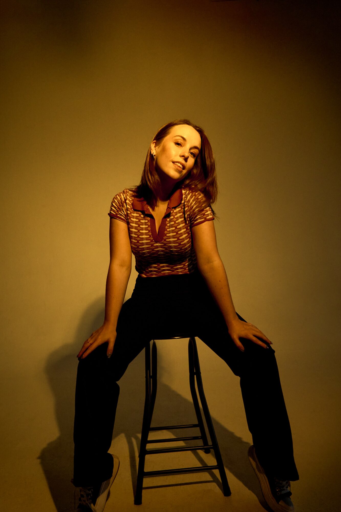 Woman sitting on a stool with hands on thighs, looking at camera, in a dimly lit room.