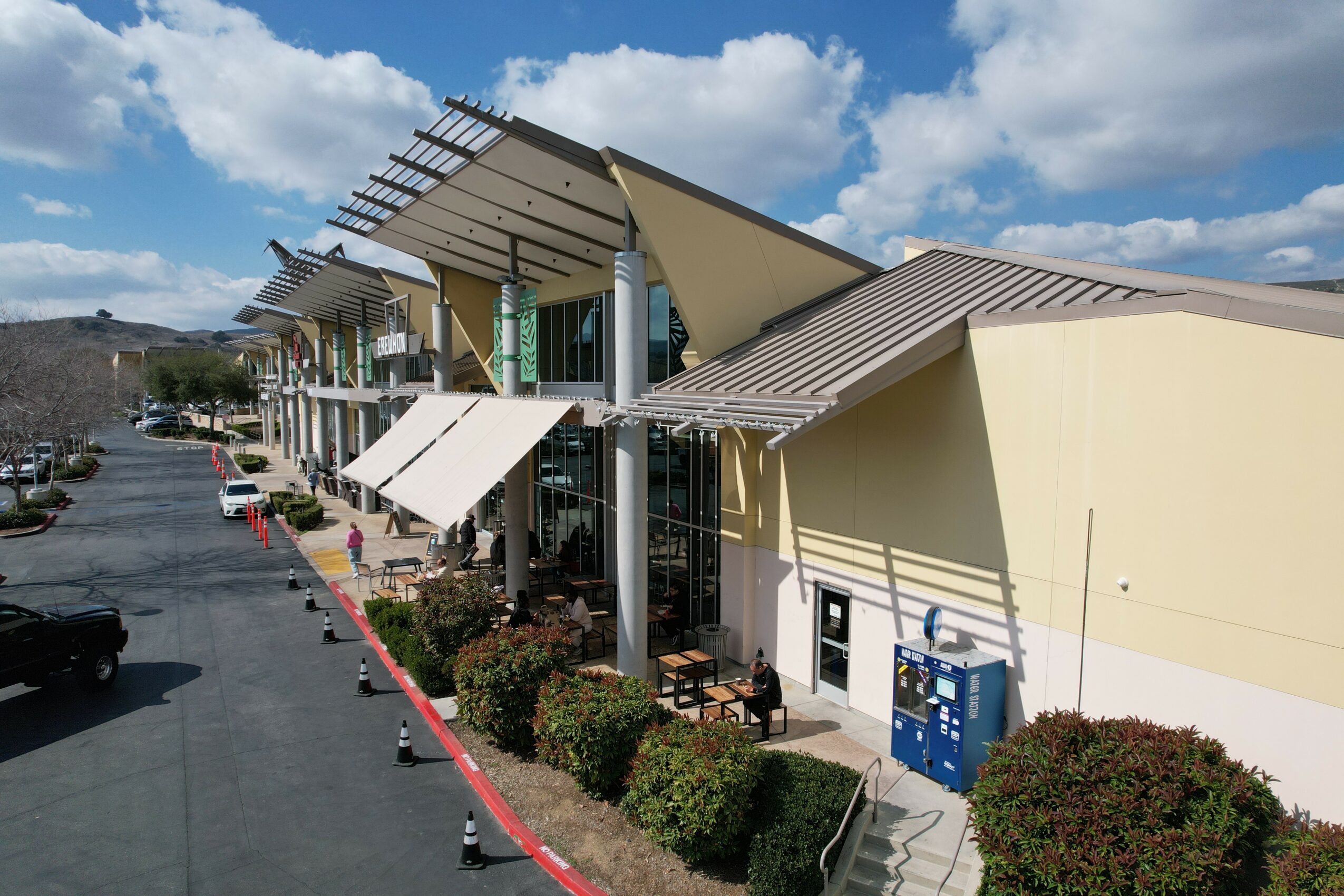 Modern building with large windows, awnings, and outdoor seating area, surrounded by bushes and parking lot, under partly cloudy sky.