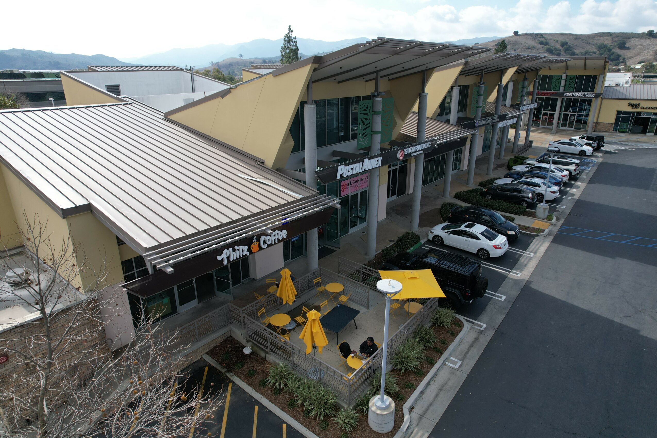 Parking lot with cars and outdoor seating area with yellow umbrellas in front of a modern building.